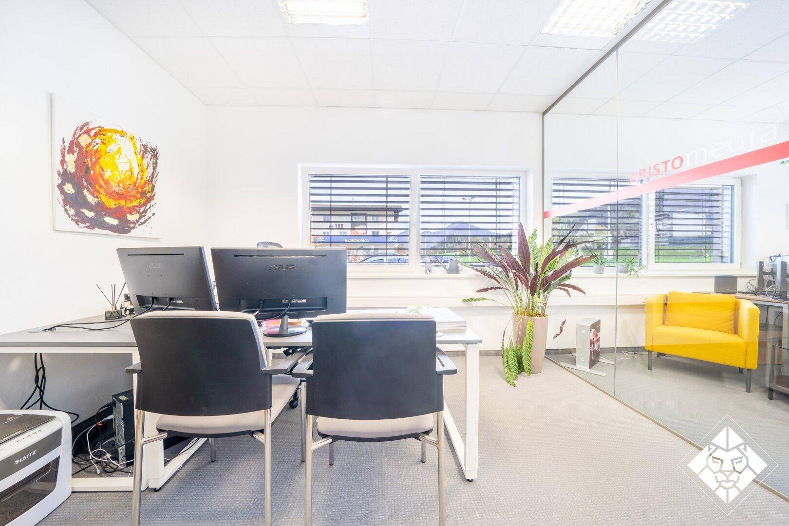 Helles Büro mit Schreibtischen, Bürostühlen und einer Glaswand zum angrenzenden Raum.