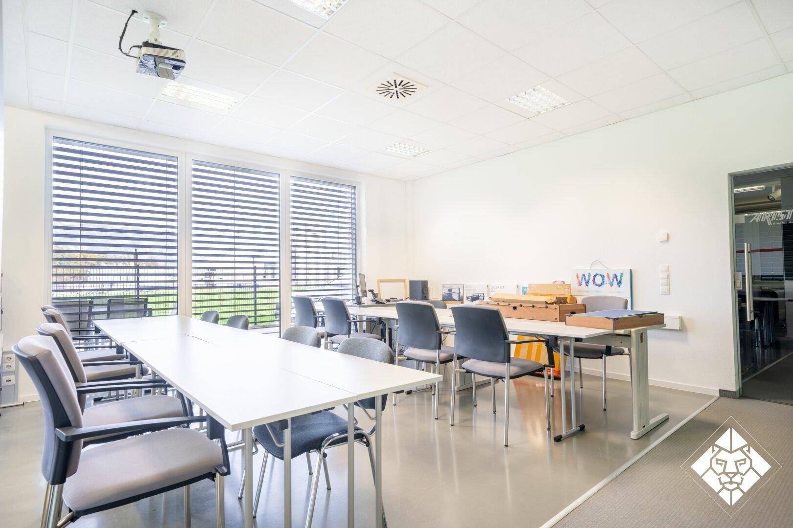 Heller Konferenzraum mit langen Tischen, Stühlen und großen Fenstern mit Jalousien.