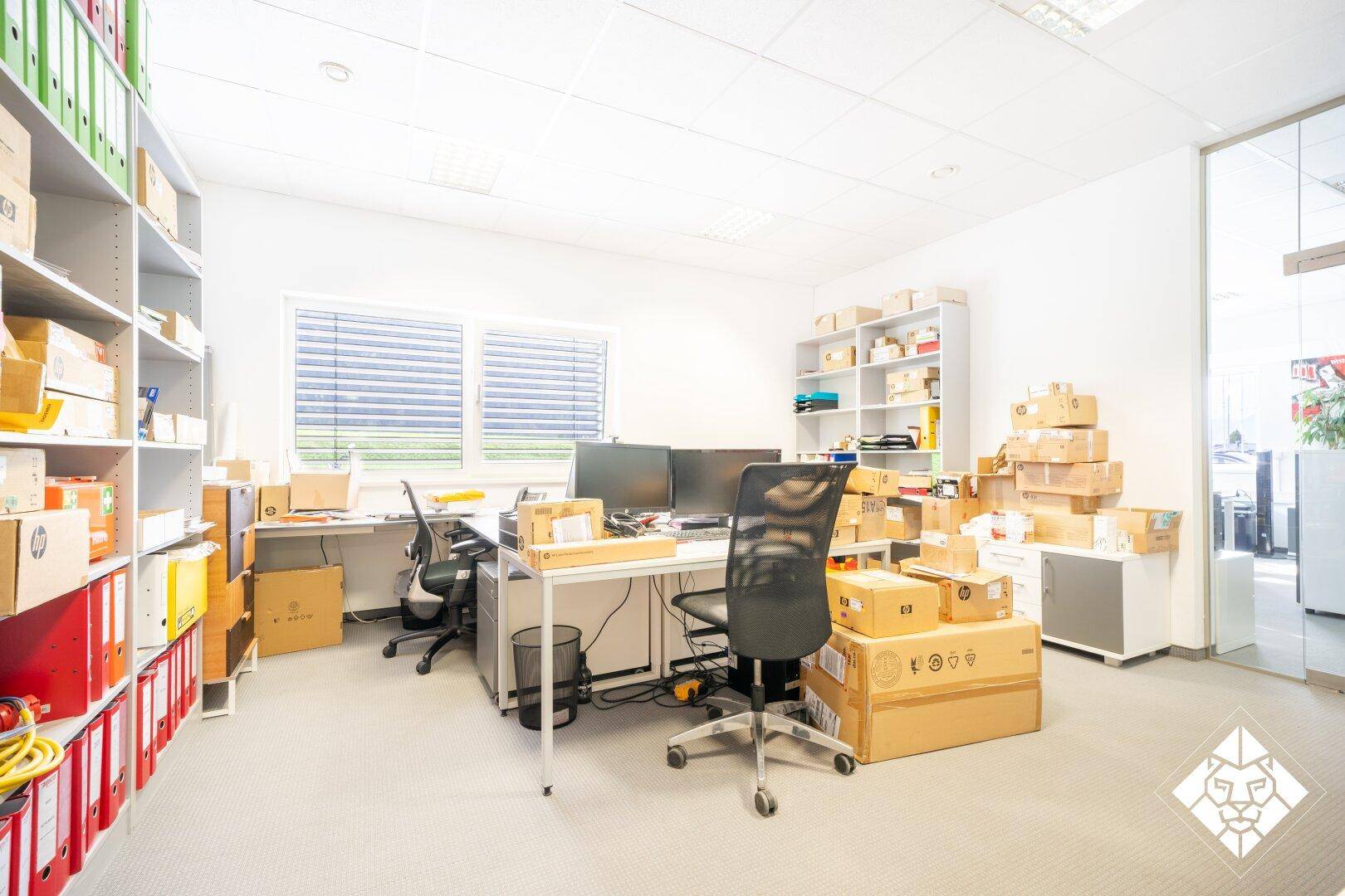 Büro mit Schreibtischen, Computern und Regalen voller Kartons und Aktenordner.
