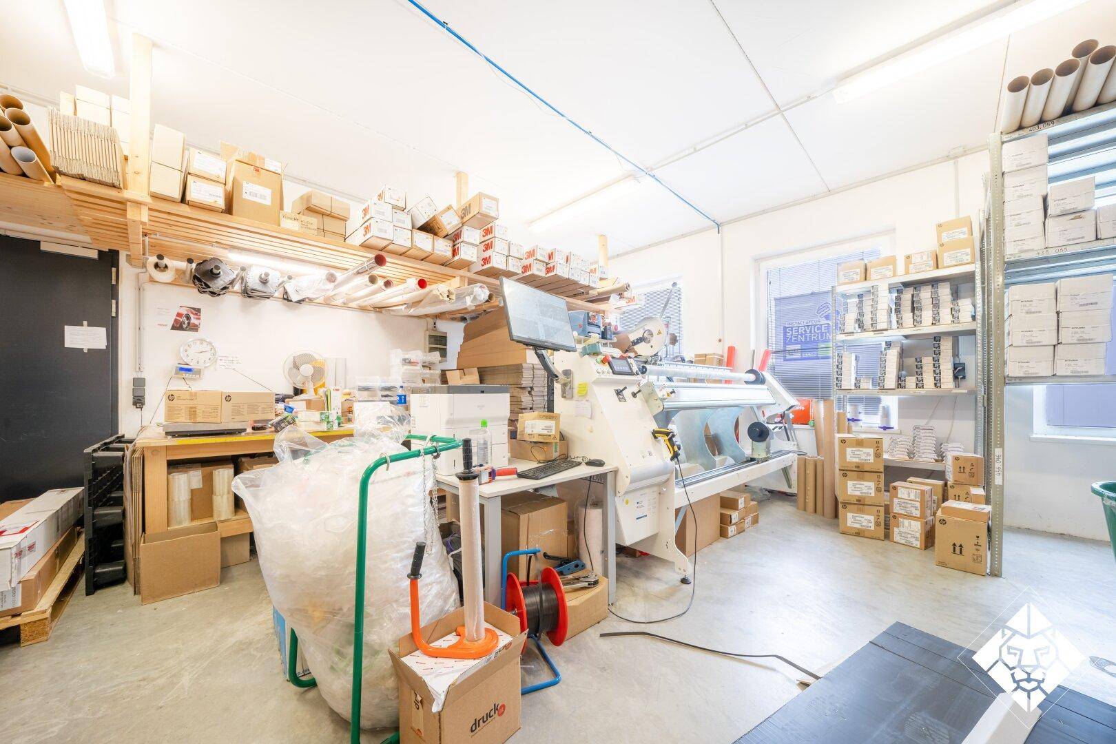 Werkstattbereich mit Arbeitsplatz, Maschinen und Regalen voller Materialien und Kartons.