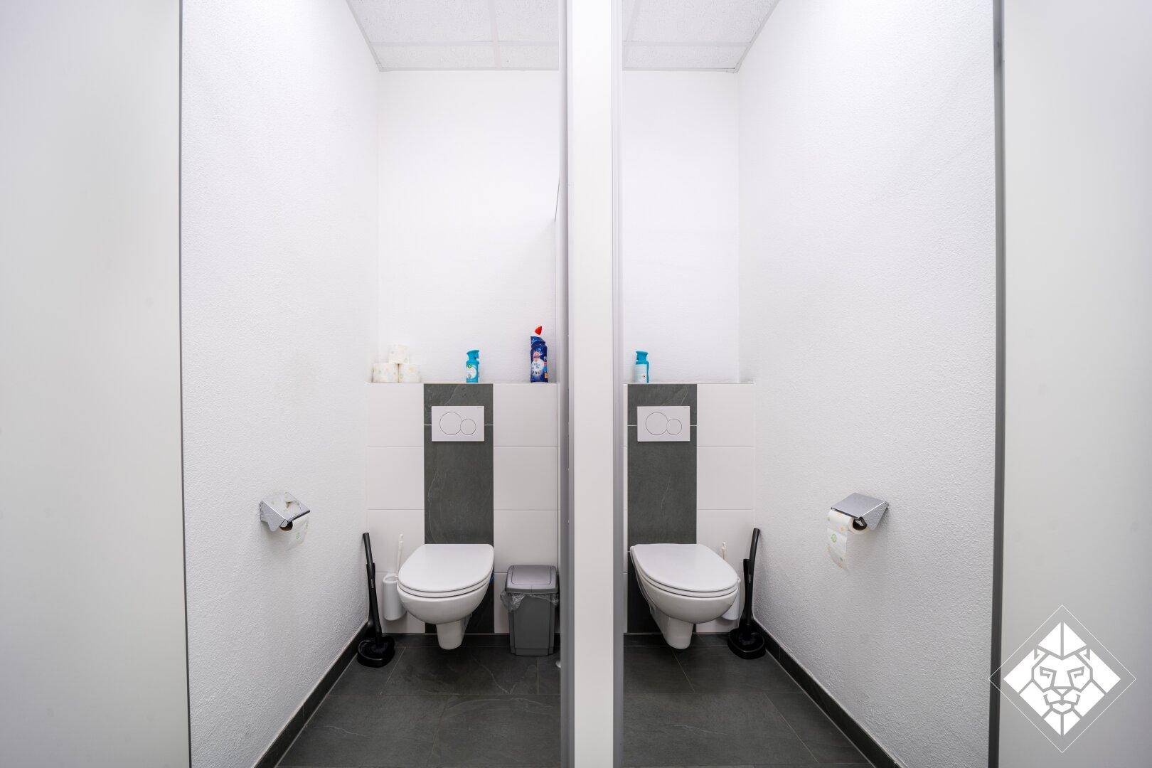 Zwei separate Toilettenkabinen mit modernen Wand-WCs und grauen Fliesenböden in einem Waschraum.