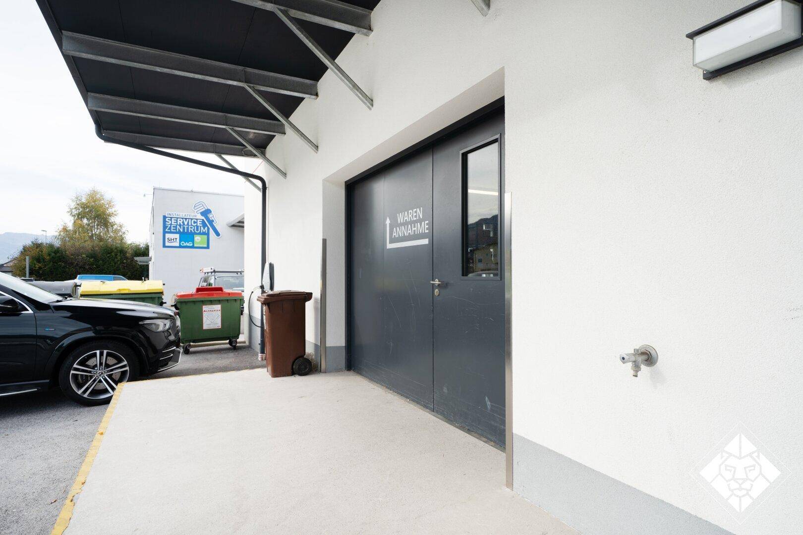 Außenansicht des Wareneingangs eines Gebäudes mit Parkplatz und Mülltonnen im Vordergrund.