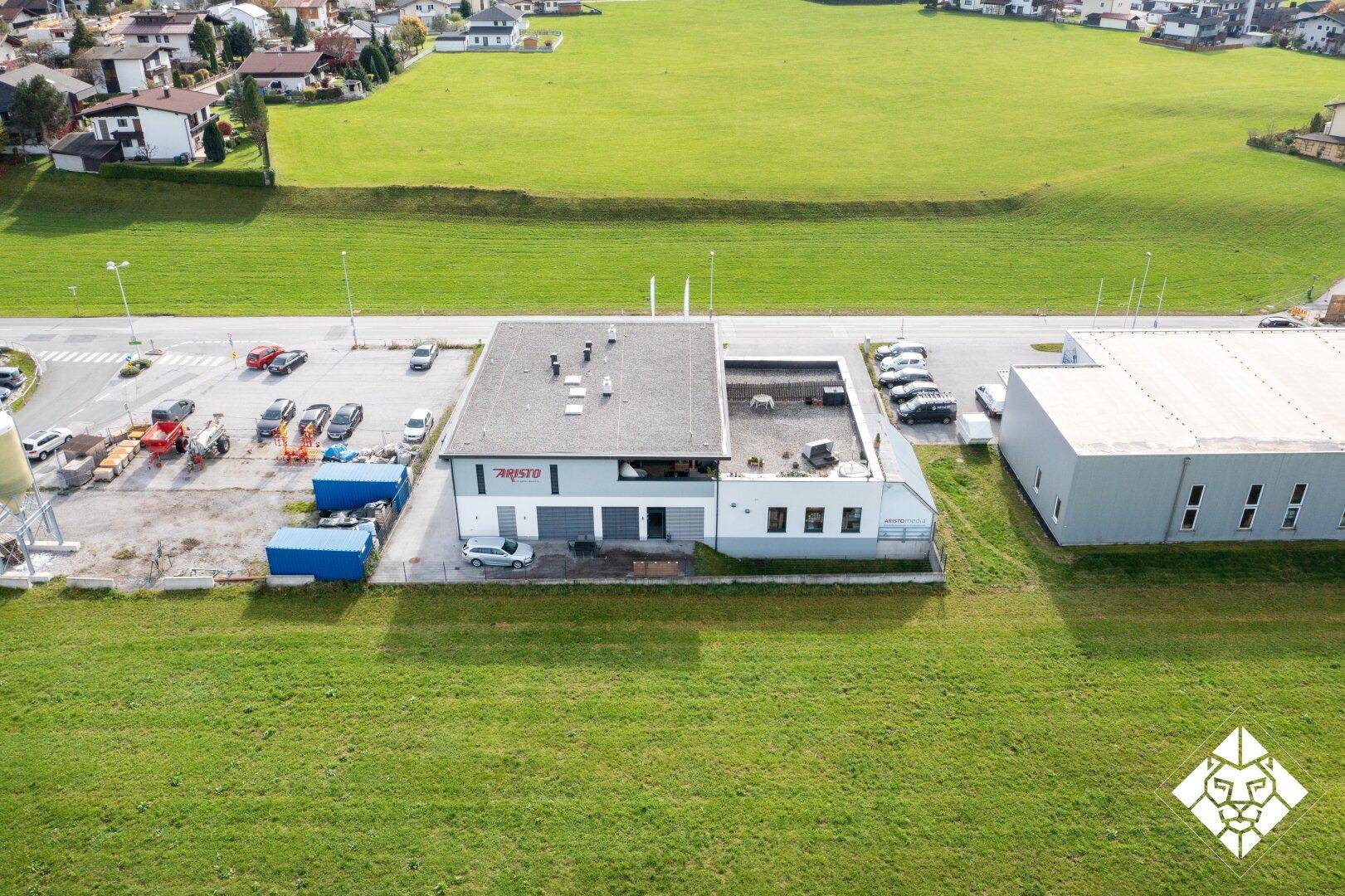 Weitläufige Luftaufnahme eines Gewerbegebäudes umgeben von grünen Feldern und Parkplätzen.