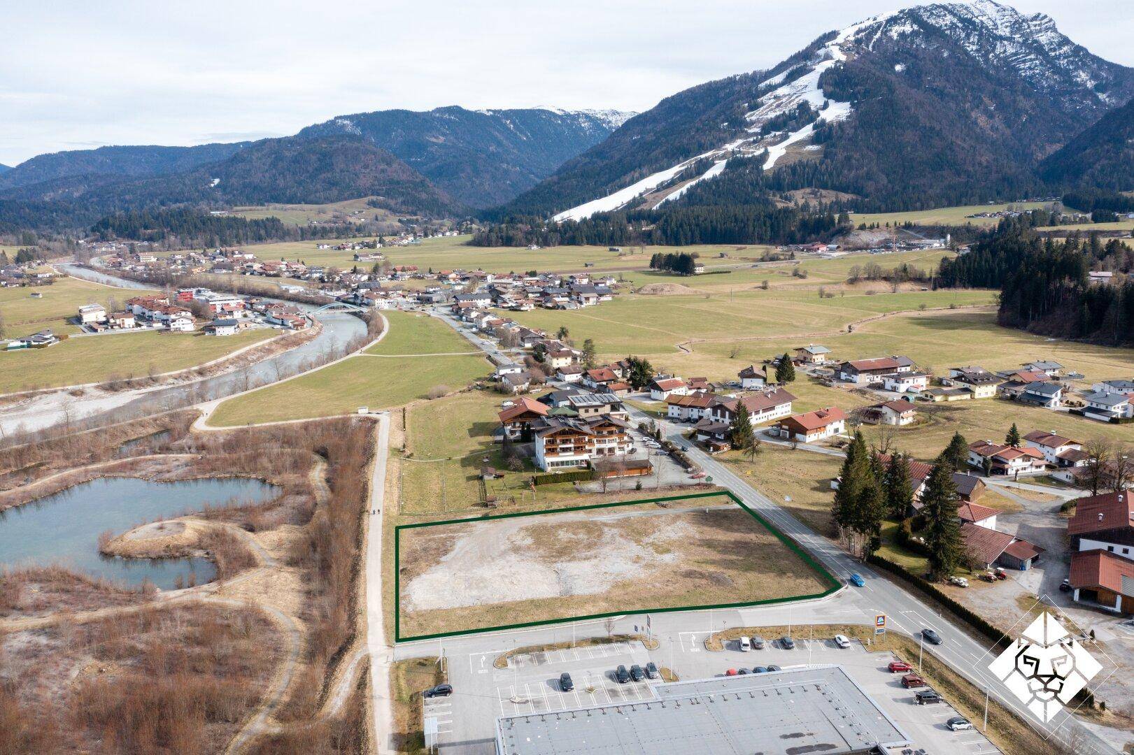 Luftaufnahme eines Baugrundstücks mit grüner Umrandung, umgeben von einem Fluss, Bergen und einer kleinen Siedlung.