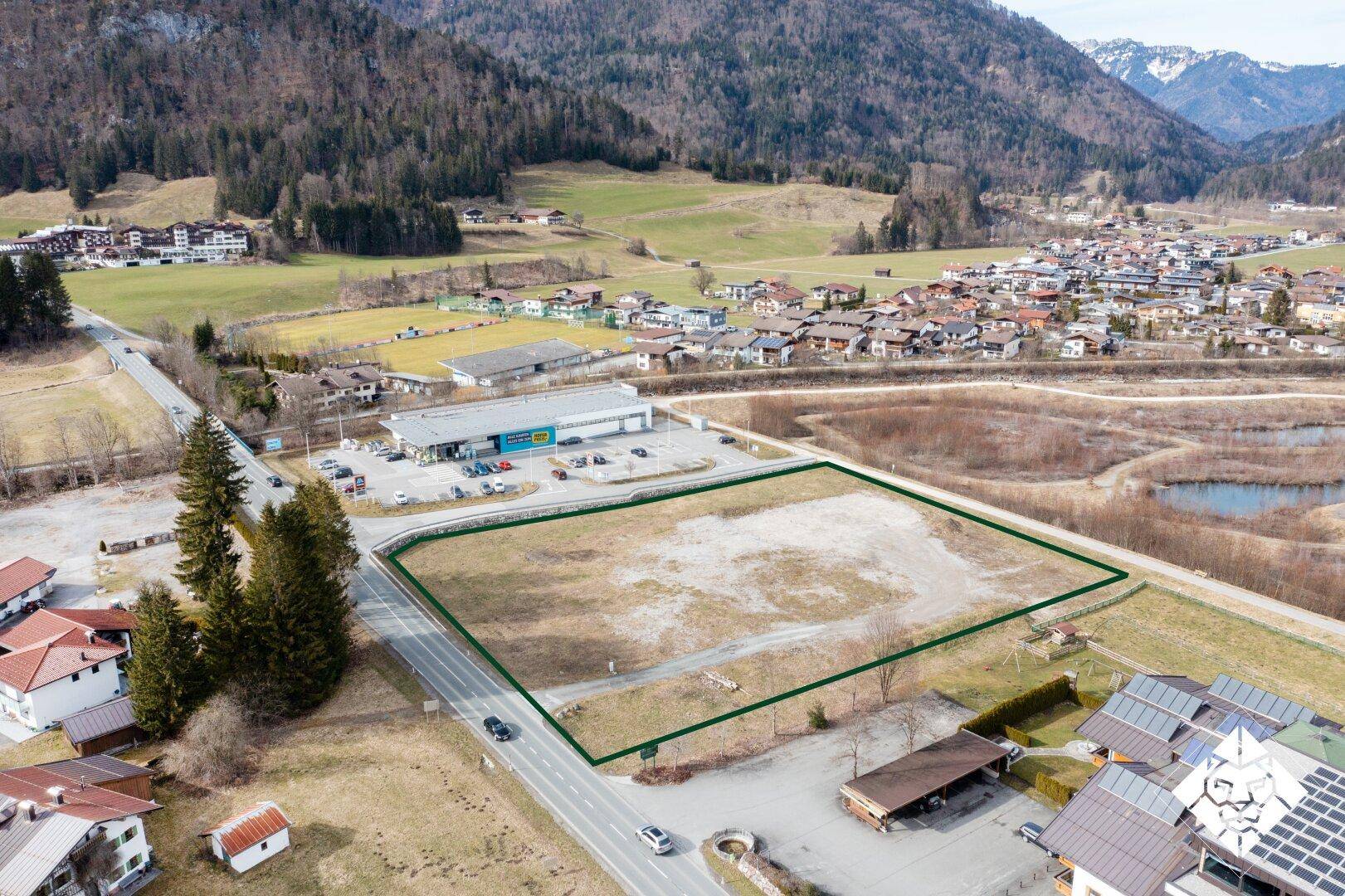 Luftbild eines Baugrundstücks mit grüner Markierung, angrenzend an einen Supermarkt und eine Landstraße.