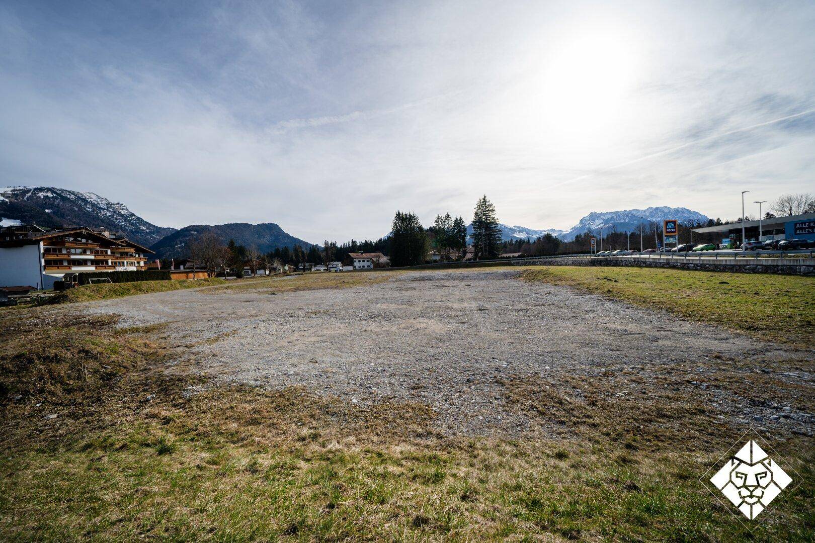 Blick über das unbebaute Grundstück mit Schotterfläche und Gras, im Hintergrund Berge und ein Supermarkt.
