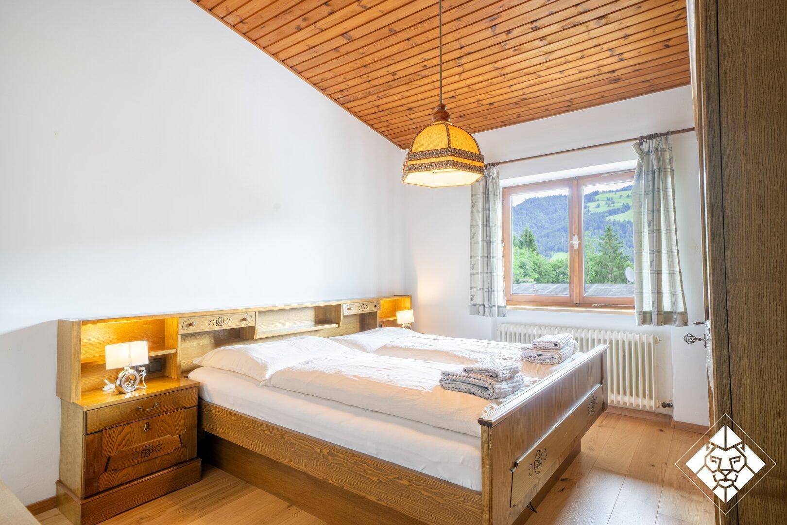Gemütliches Schlafzimmer mit Holzbett, Holzdecke und Fenster mit Blick auf die grüne Berglandschaft.