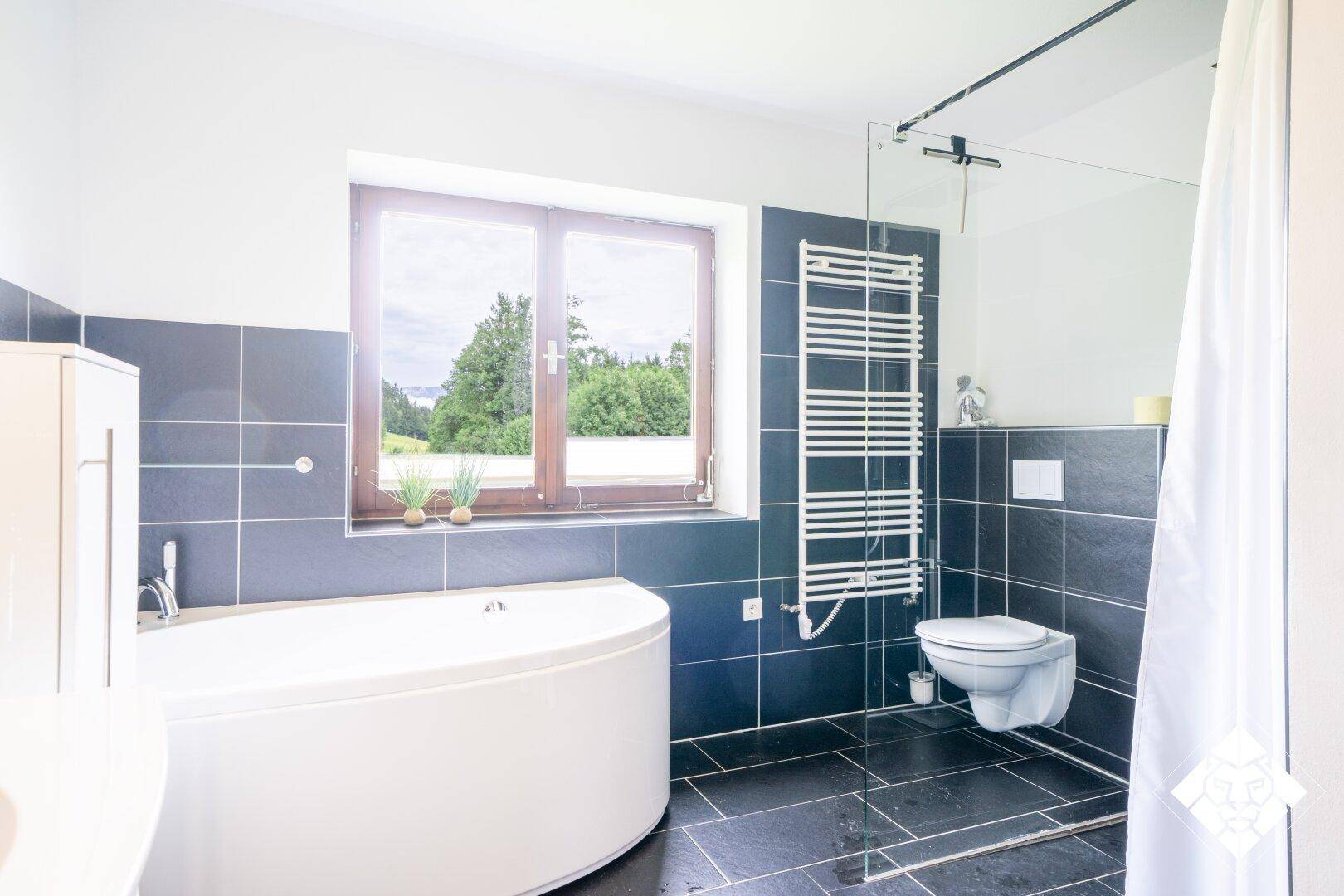 Modernes Badezimmer mit dunklen Fliesen, Eckbadewanne, Wand-WC und Fenster mit schöner Aussicht.