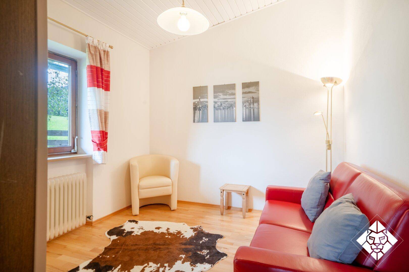 Helles Wohnzimmer mit rotem Sofa, Sessel und Holzboden, ergänzt durch einen Kuhfellteppich.