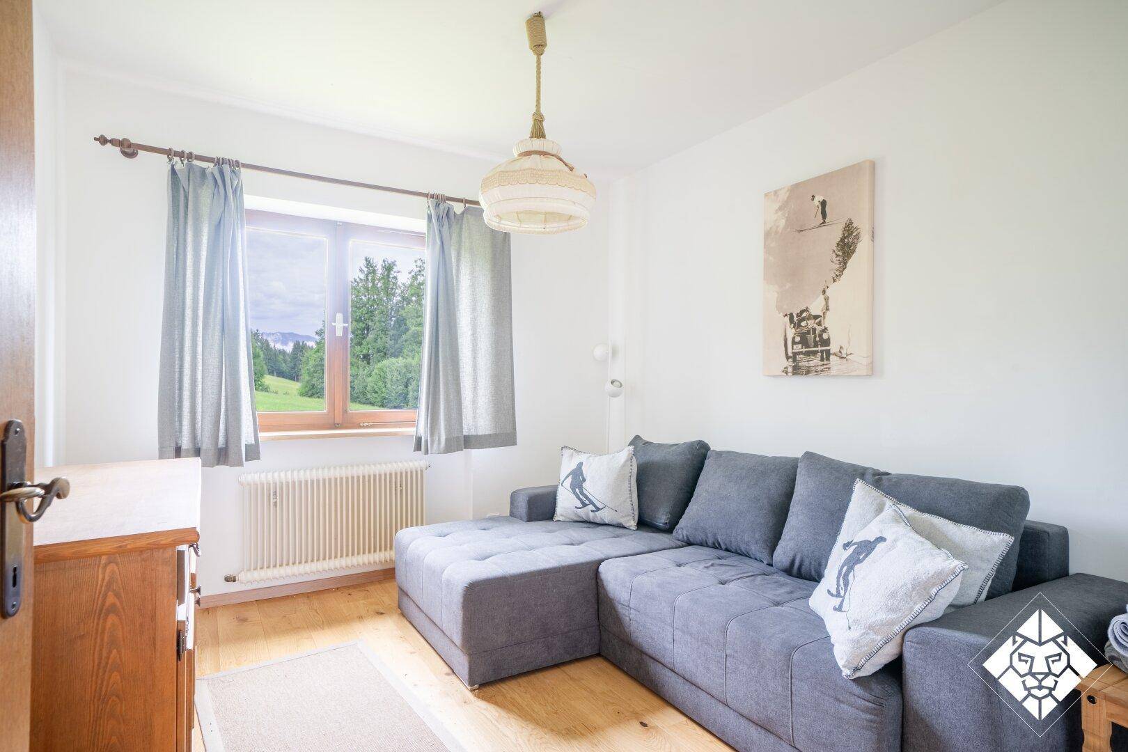 Gemütliches Wohnzimmer mit grauem Ecksofa, Holzboden und Fenster mit Blick ins Grüne.