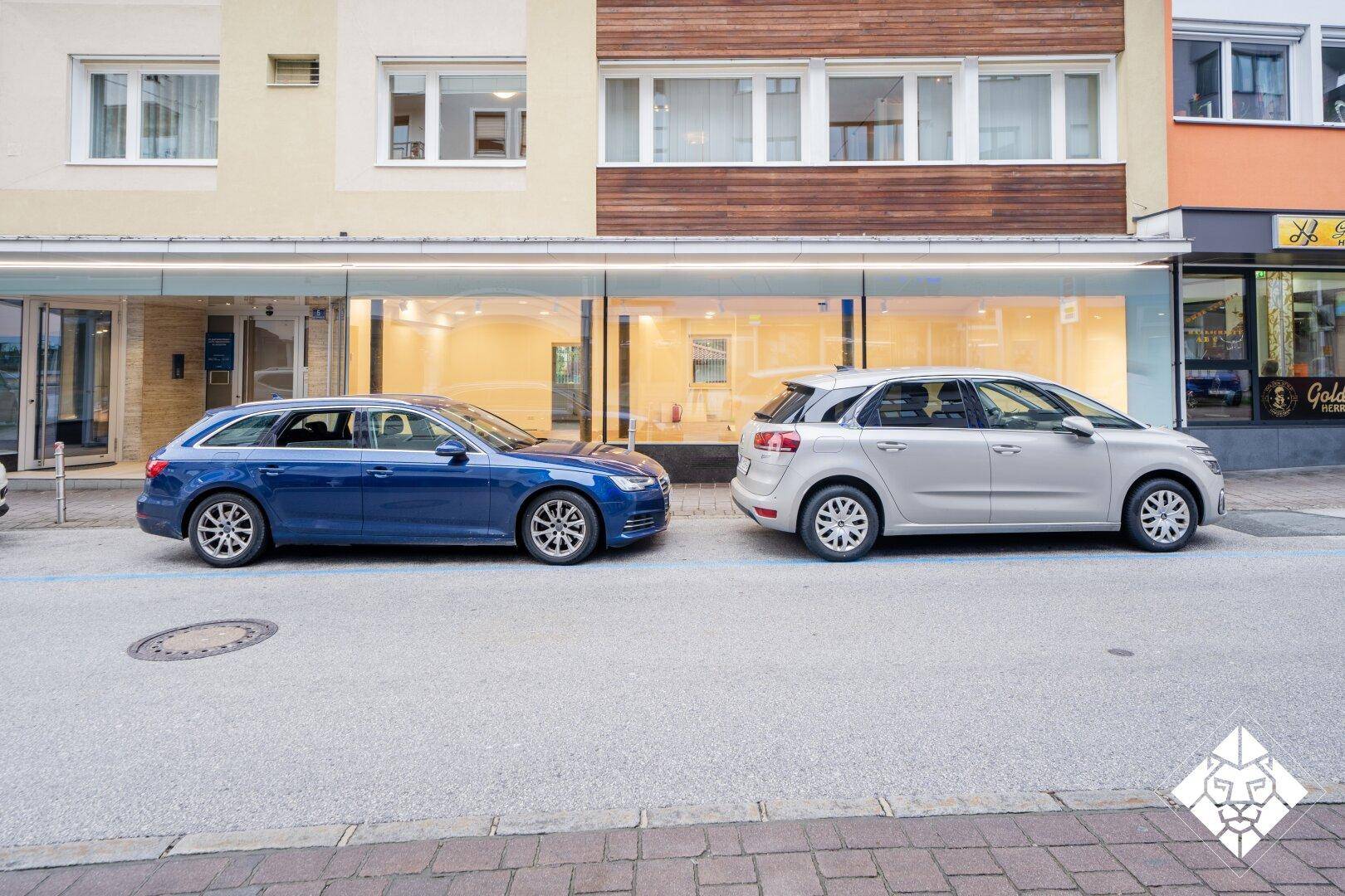 Gebäudefassade mit großer Glasfront im Erdgeschoss und darüber liegenden Wohnungen an einer belebten Straße.
