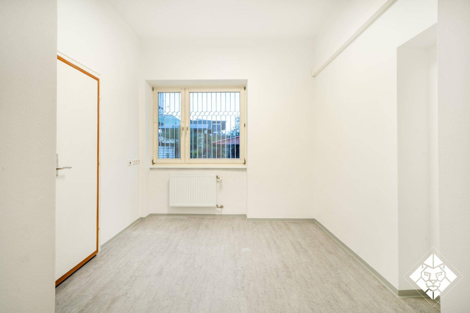 Hinterer Raum mit hellem Boden, Fenster mit Gitter, Heizkörper und weißer Tür mit Holzrahmen.