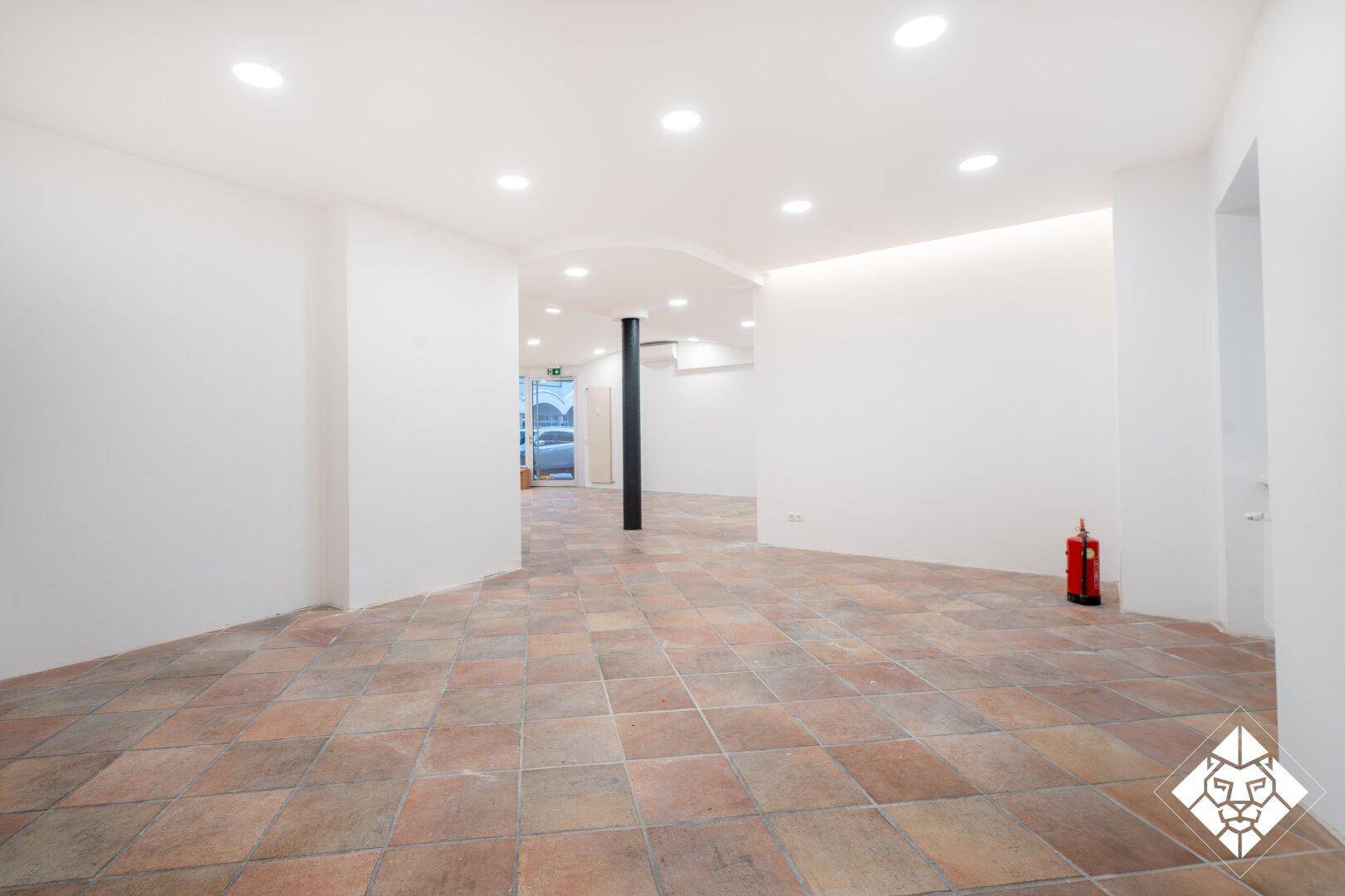 Übergangsbereich im Geschäftsraum mit Terrakotta-Fliesenboden, weiß gestrichenen Wänden und Deckenleuchten.