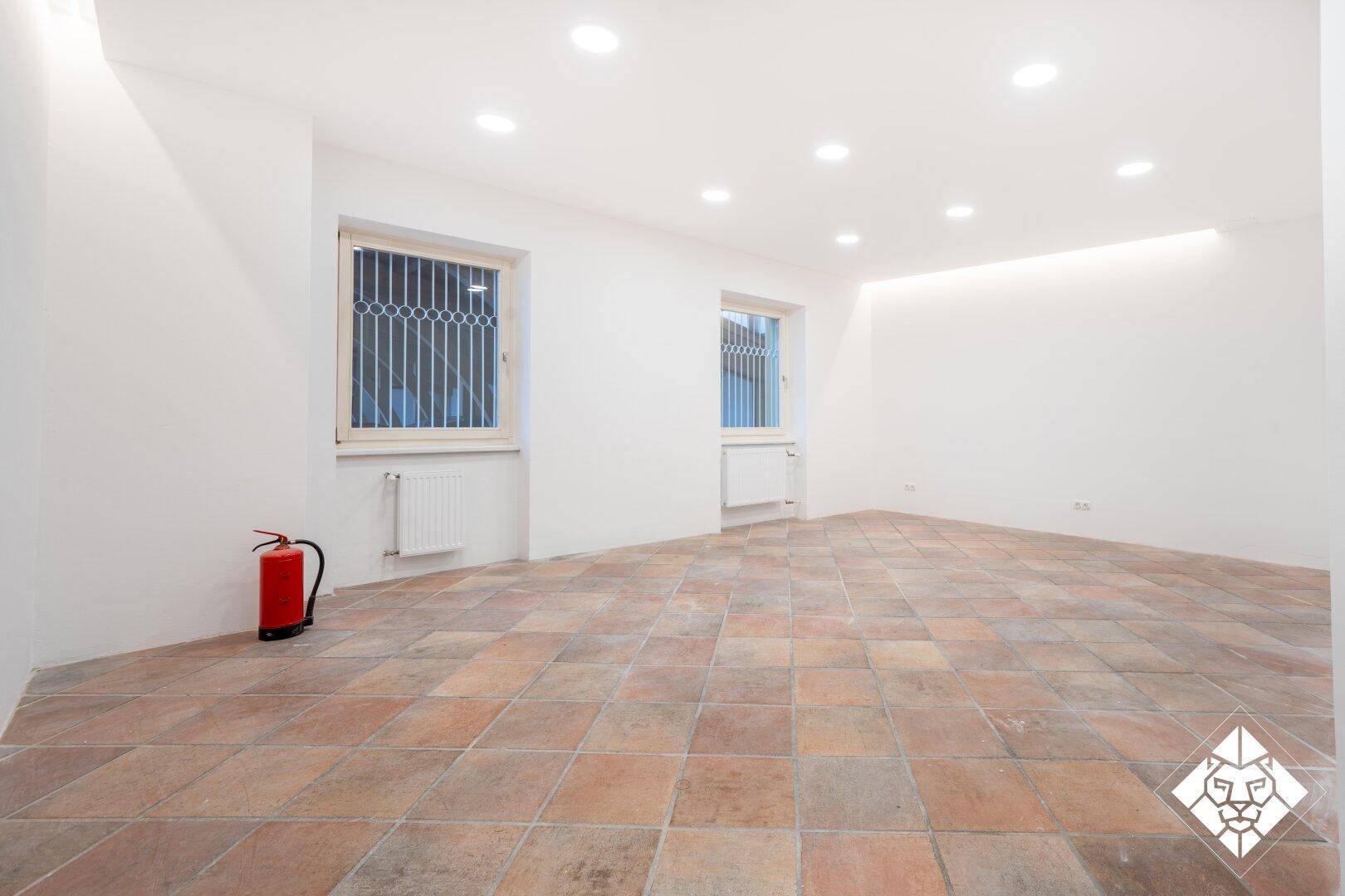 Kleinerer Raum mit Terrakotta-Fliesenboden, zwei Fenstern mit Gittern und Heizkörpern.