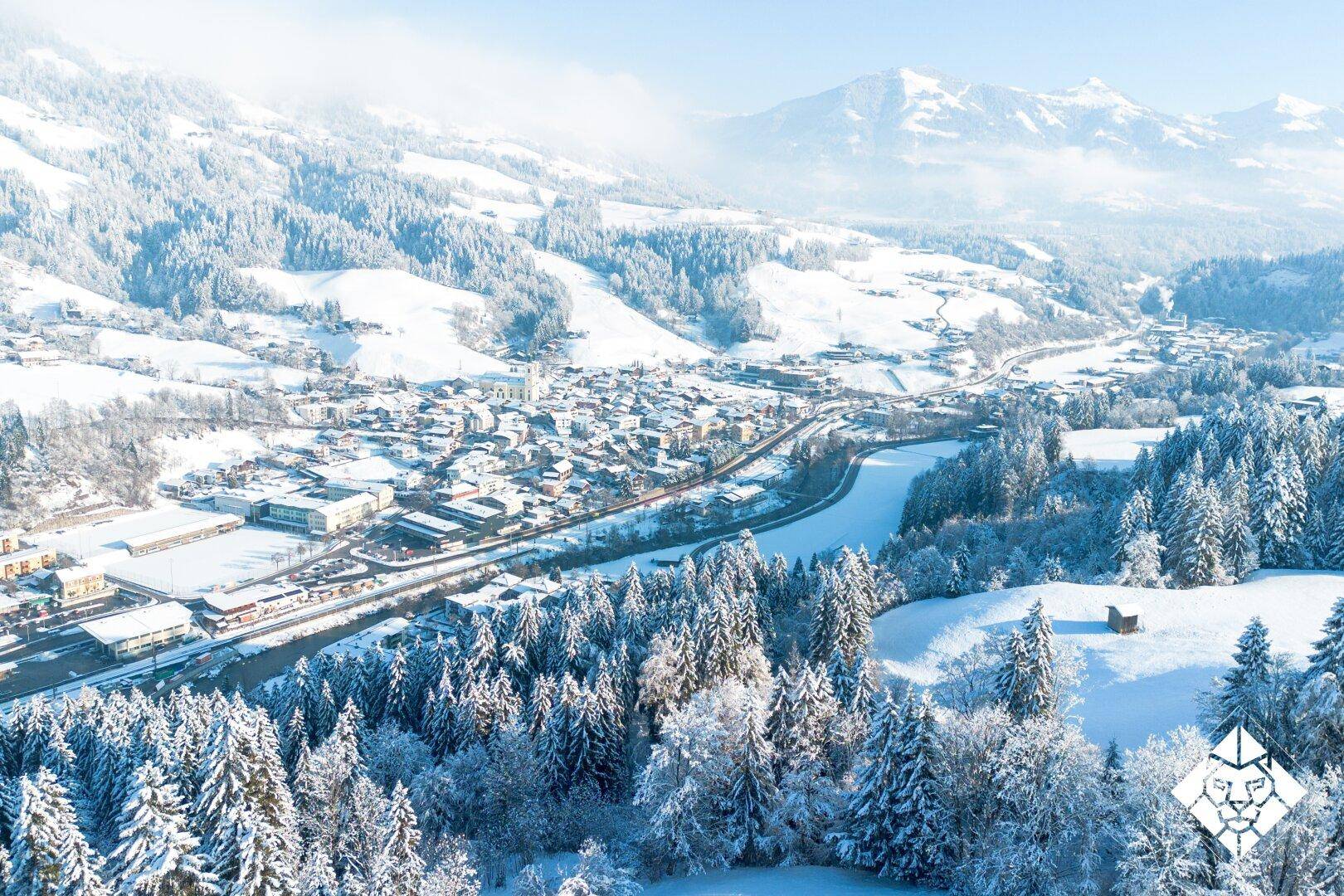 Weitläufige Winterlandschaft mit verschneiter Stadt, Wäldern und majestätischen Bergen im Hintergrund.
