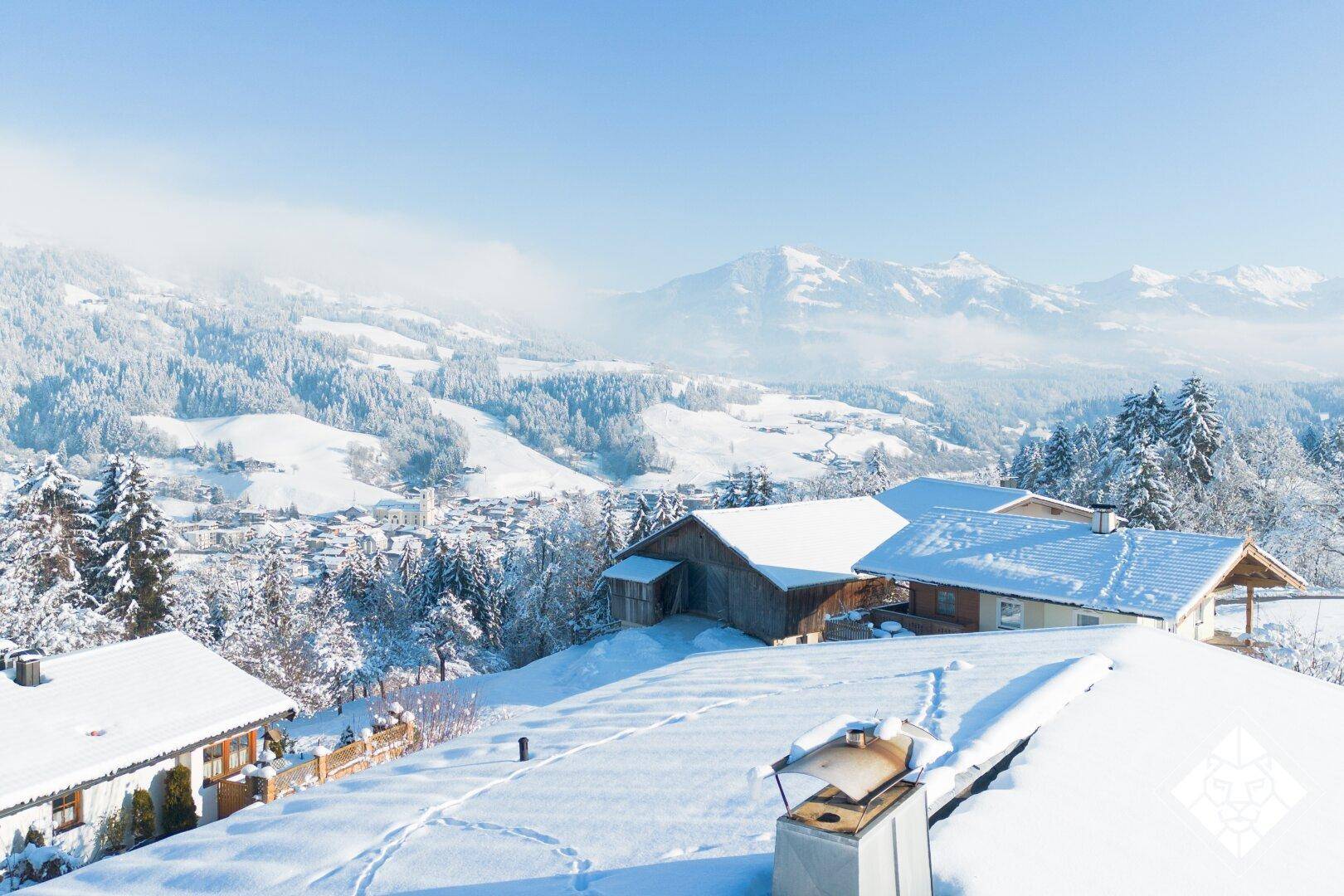 Blick über verschneite Dächer auf die umliegende Berglandschaft im Winter mit klarem Himmel.