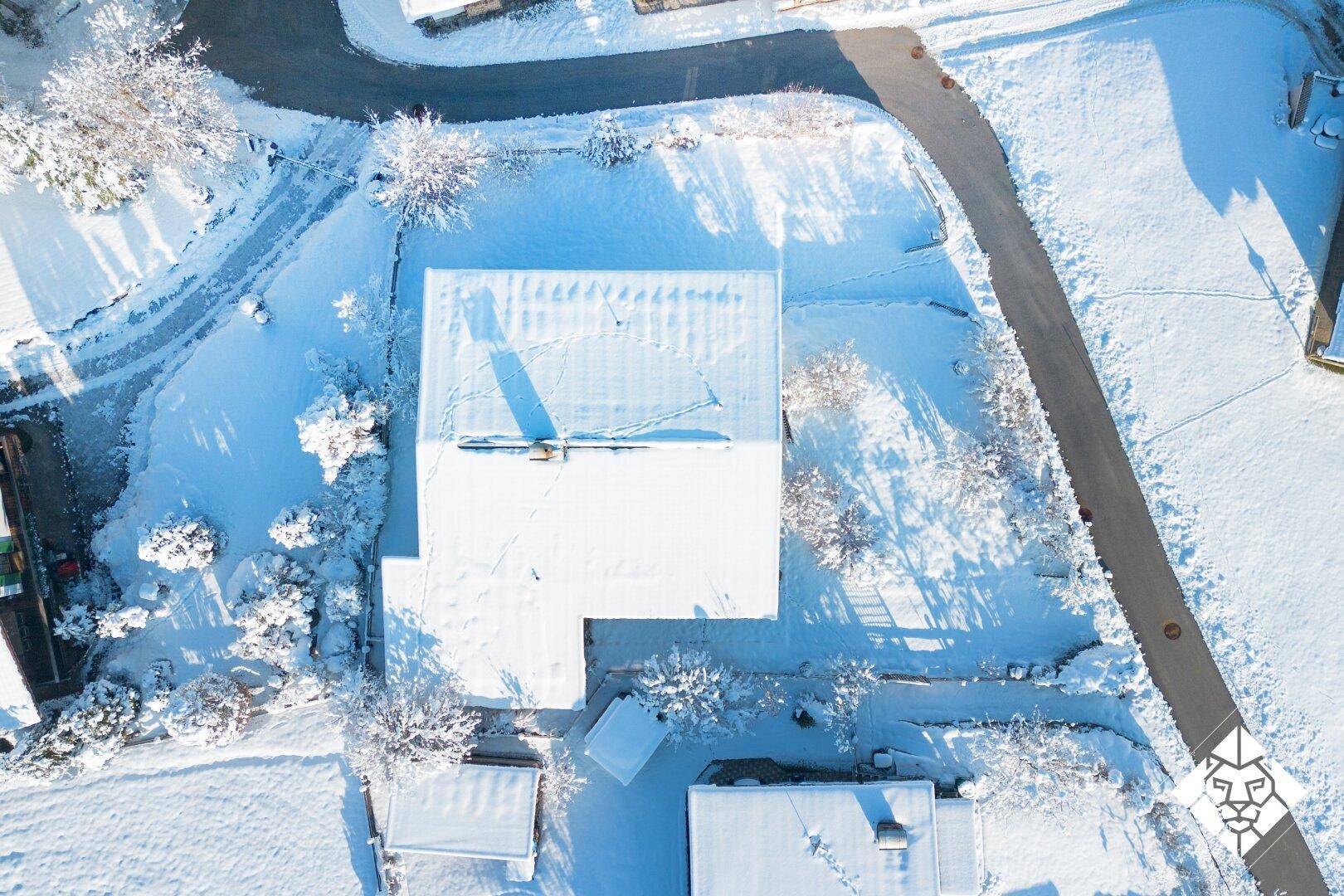 Draufsicht auf ein schneebedecktes Haus mit Zufahrt und umliegenden Bäumen in winterlicher Pracht.