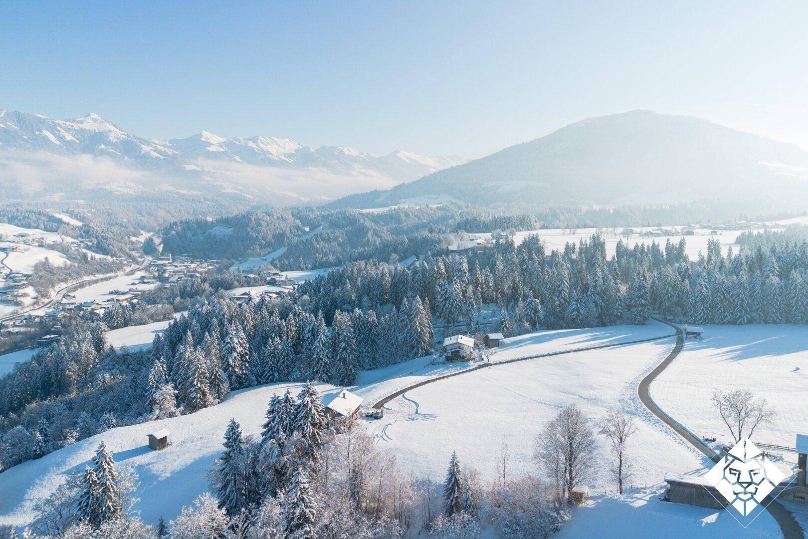 Weitläufige Winterlandschaft aus der Vogelperspektive mit verschneiten Hügeln, Wäldern und majestätischen Bergen.