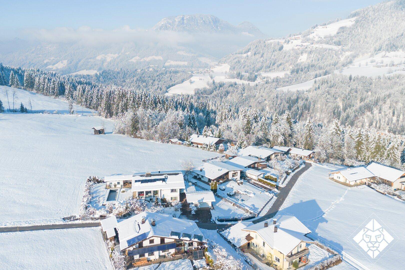 Luftaufnahme einer Siedlung in verschneiter Berglandschaft mit mehreren Häusern und Wäldern.