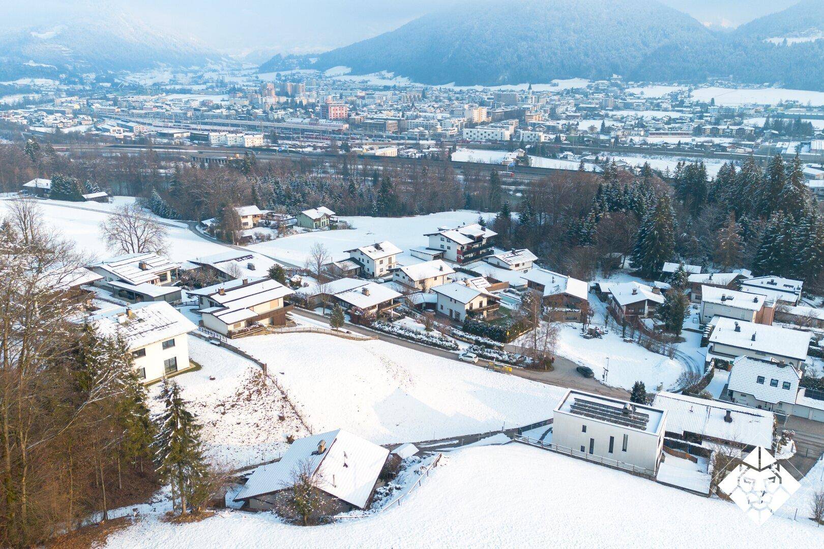 Luftaufnahme einer verschneiten Wohnsiedlung mit Bergen im Hintergrund und einer Stadt in der Ferne.