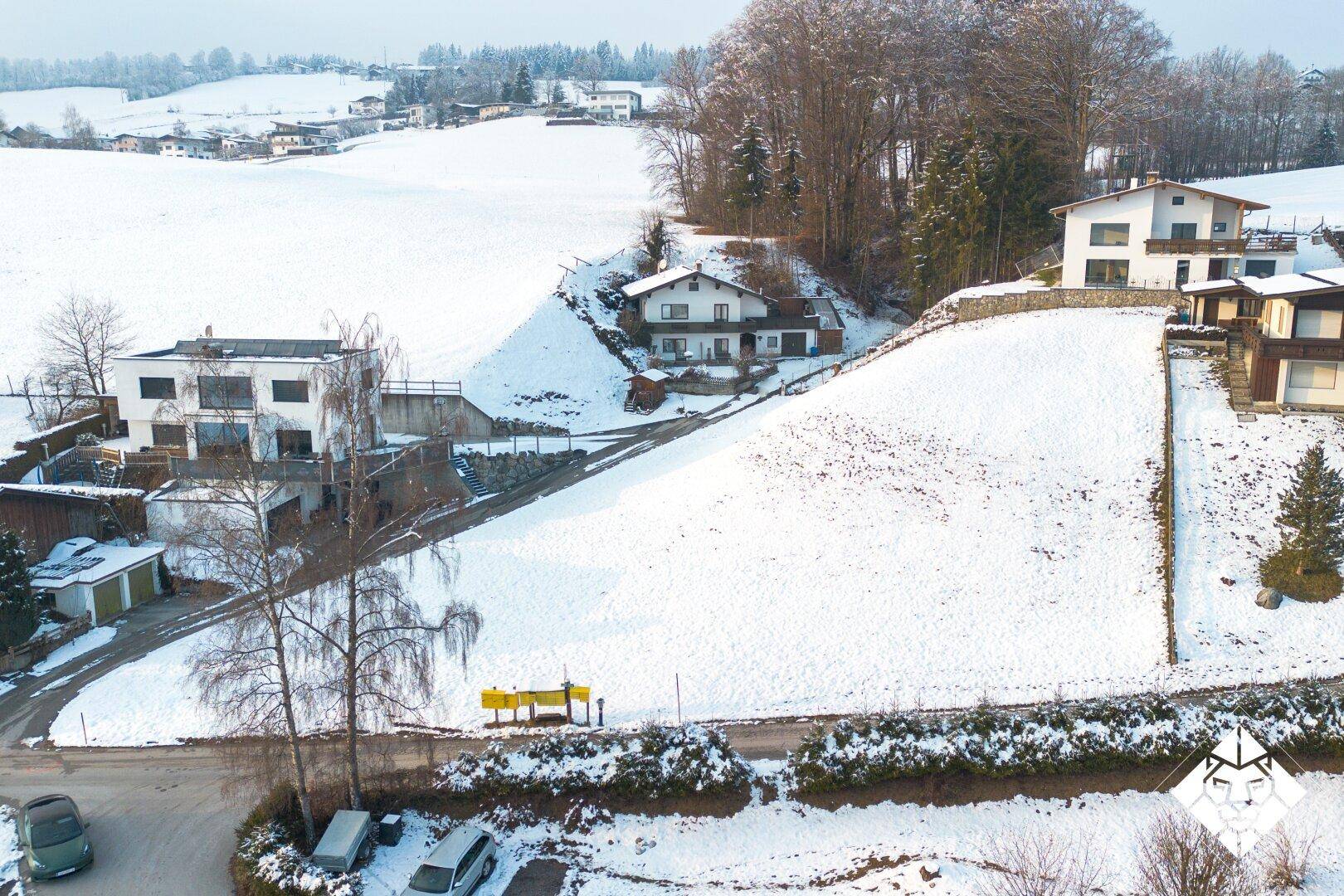 Blick aus der Vogelperspektive auf eine verschneite Wohngegend mit Häusern am Hang und einer Straße.
