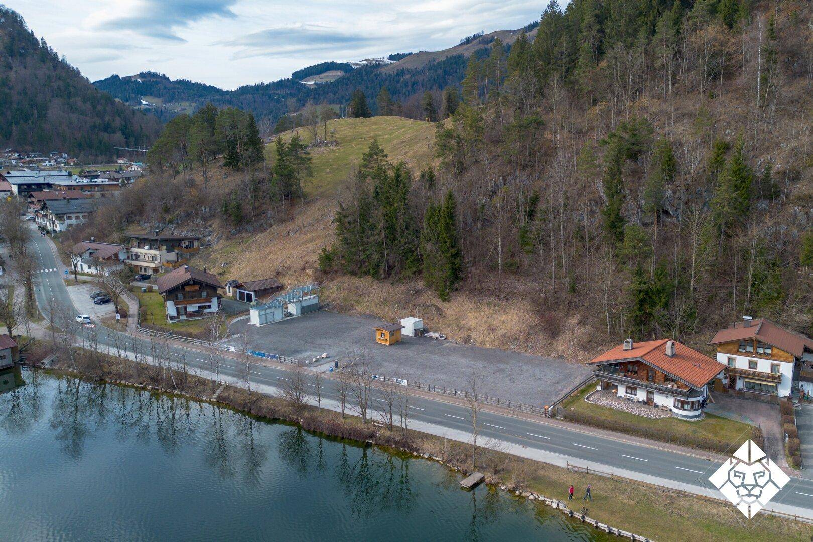 Luftaufnahme des Grundstücksbereichs mit See, Straße, umliegenden Häusern und bewaldetem Hang.
