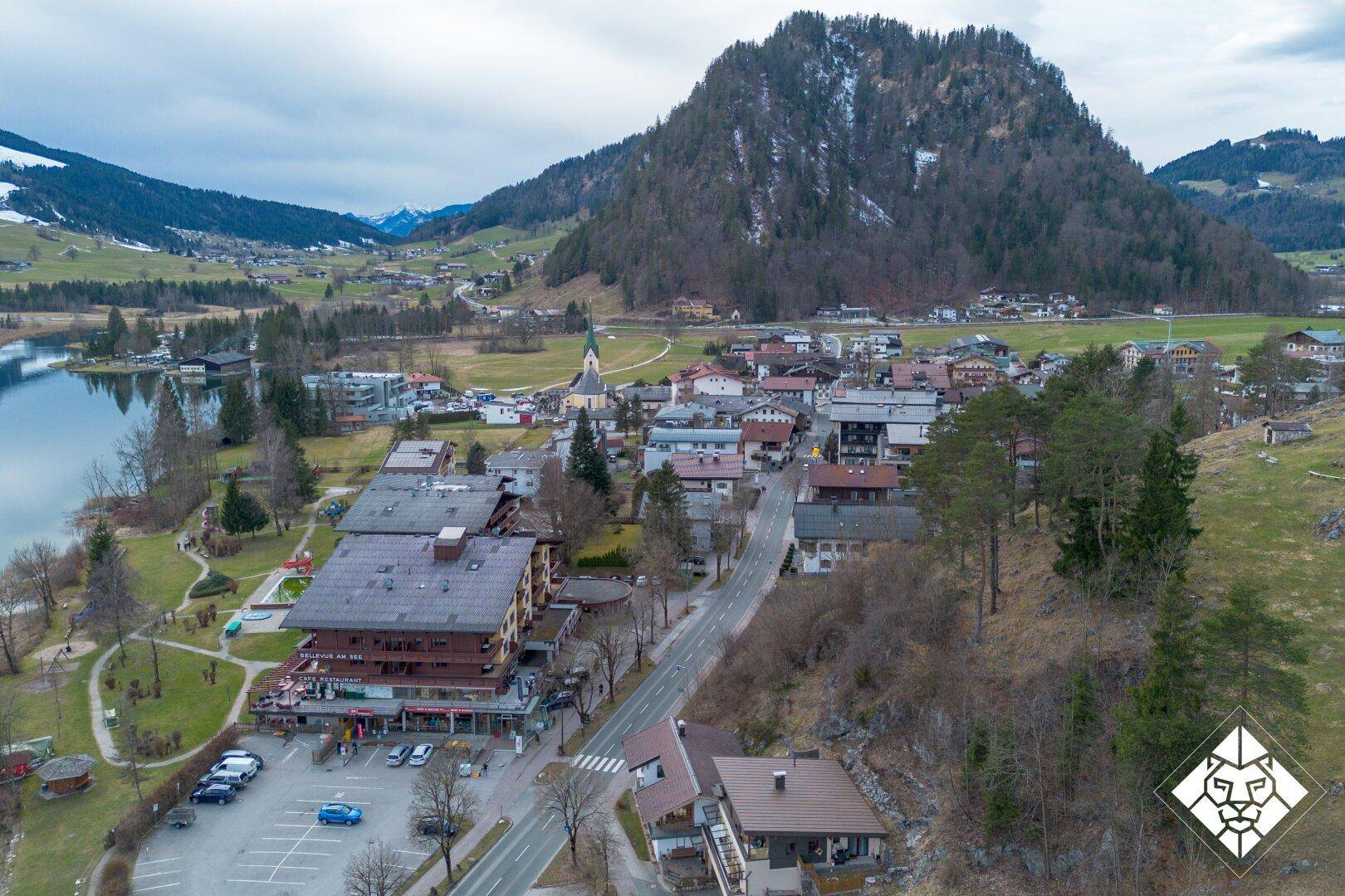 Dorfansicht am Seeufer mit traditionellen Gebäuden und bewaldeten Bergen im Hintergrund.