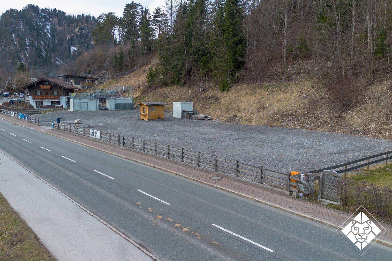 Grundstück mit Schotterfläche neben einer Straße und bewaldetem Hang im Hintergrund.