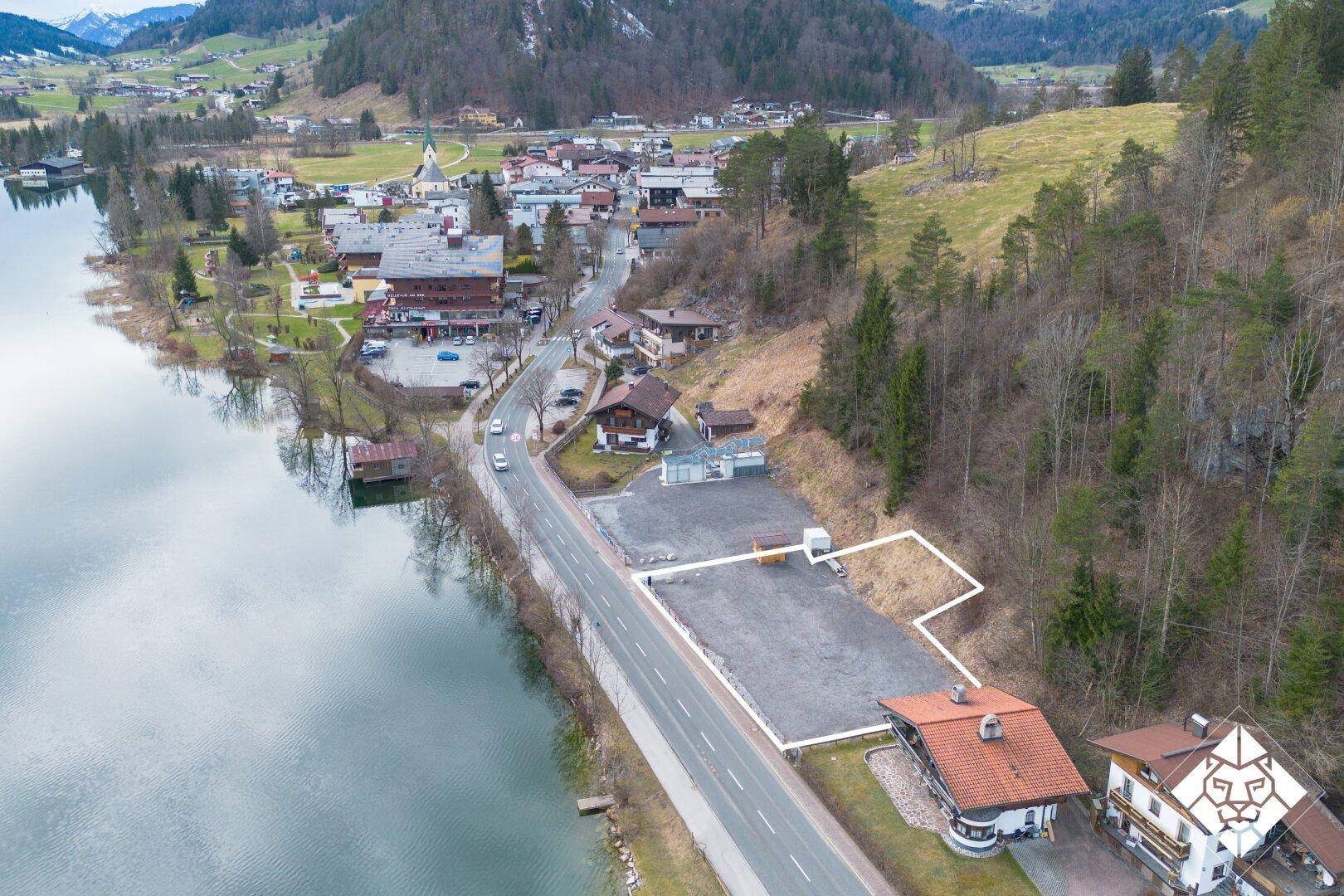 Luftaufnahme eines Grundstücks am Seeufer mit umliegender Bebauung und Bergen im Hintergrund.