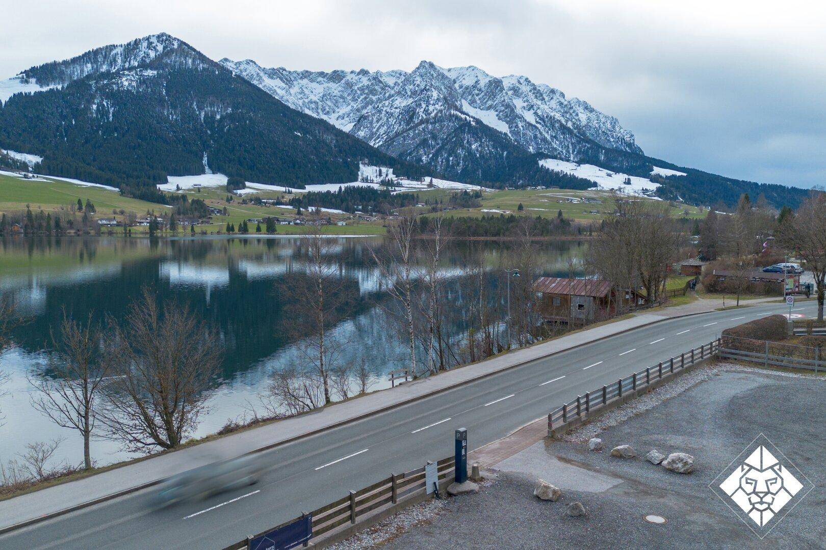 Blick von der Straße über den See auf die beeindruckende Kulisse schneebedeckter Berge.