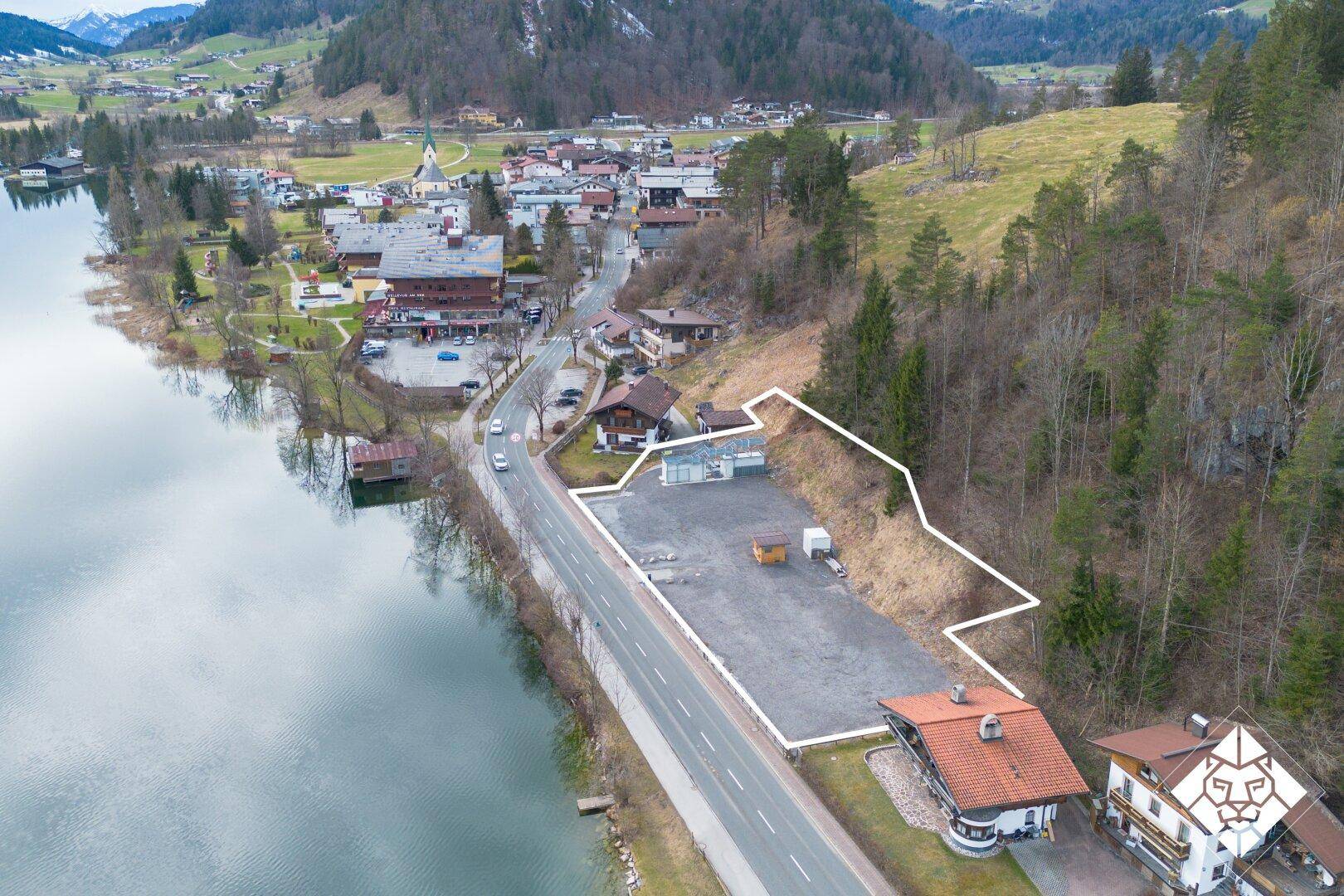 Luftaufnahme eines Grundstücks am Seeufer mit umliegenden Gebäuden und Berglandschaft.