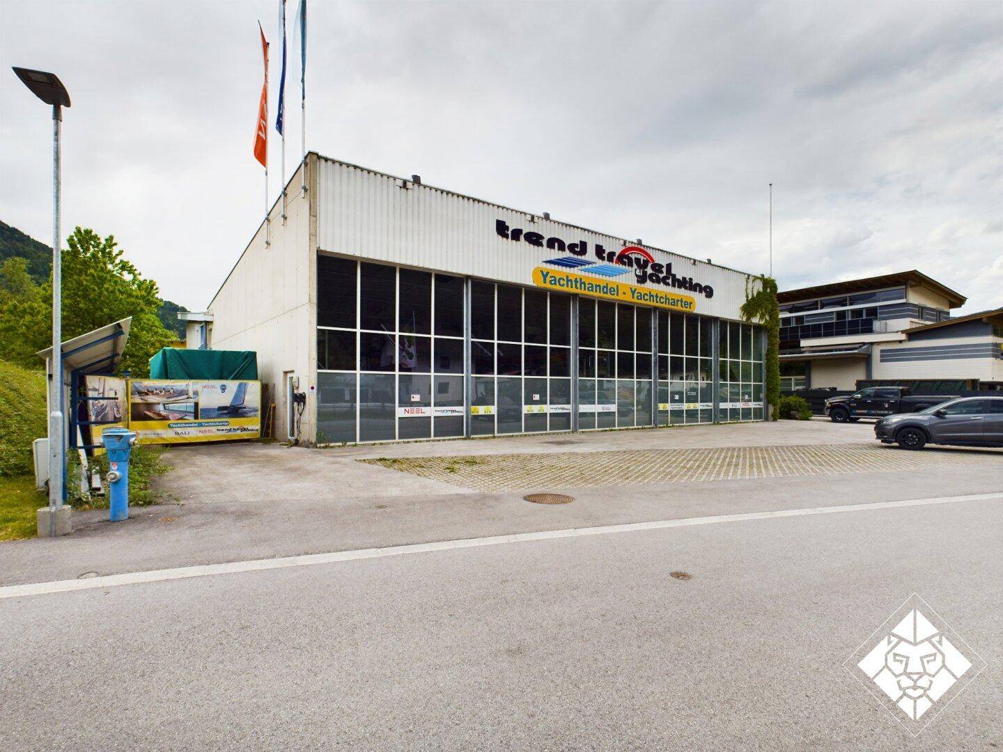 Gewerbegebäude mit großer Glasfront und Parkplätzen, umgeben von Bäumen und einer Berglandschaft.