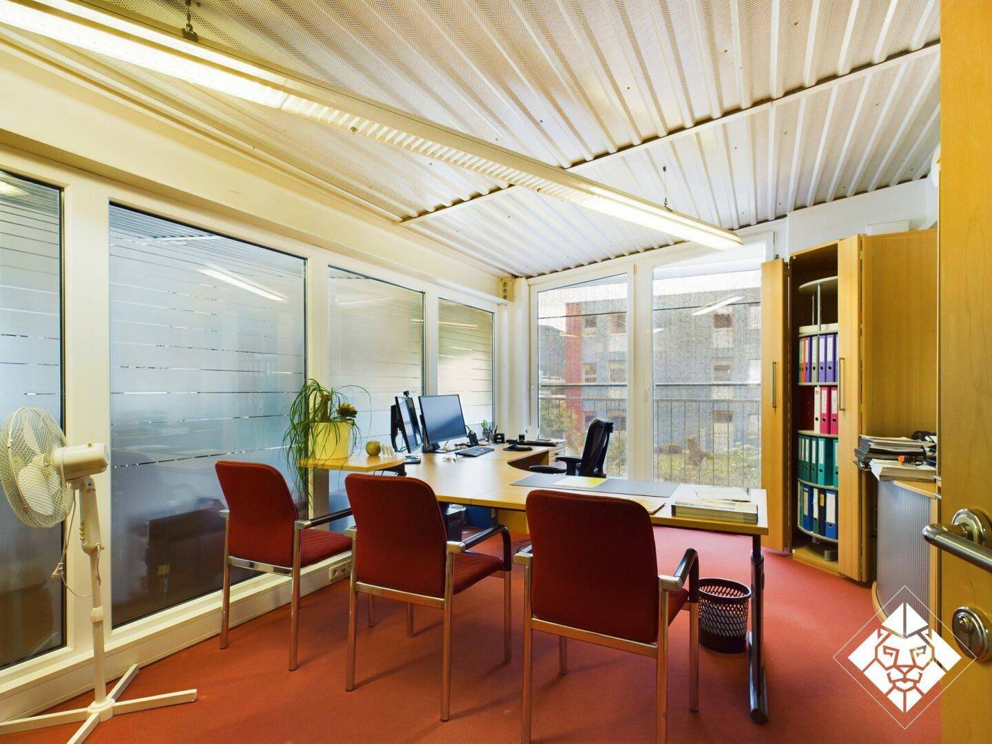 Einzelbüro mit rotem Teppichboden, Schreibtisch, Stühlen und großen Fenstern mit Blick nach draußen.