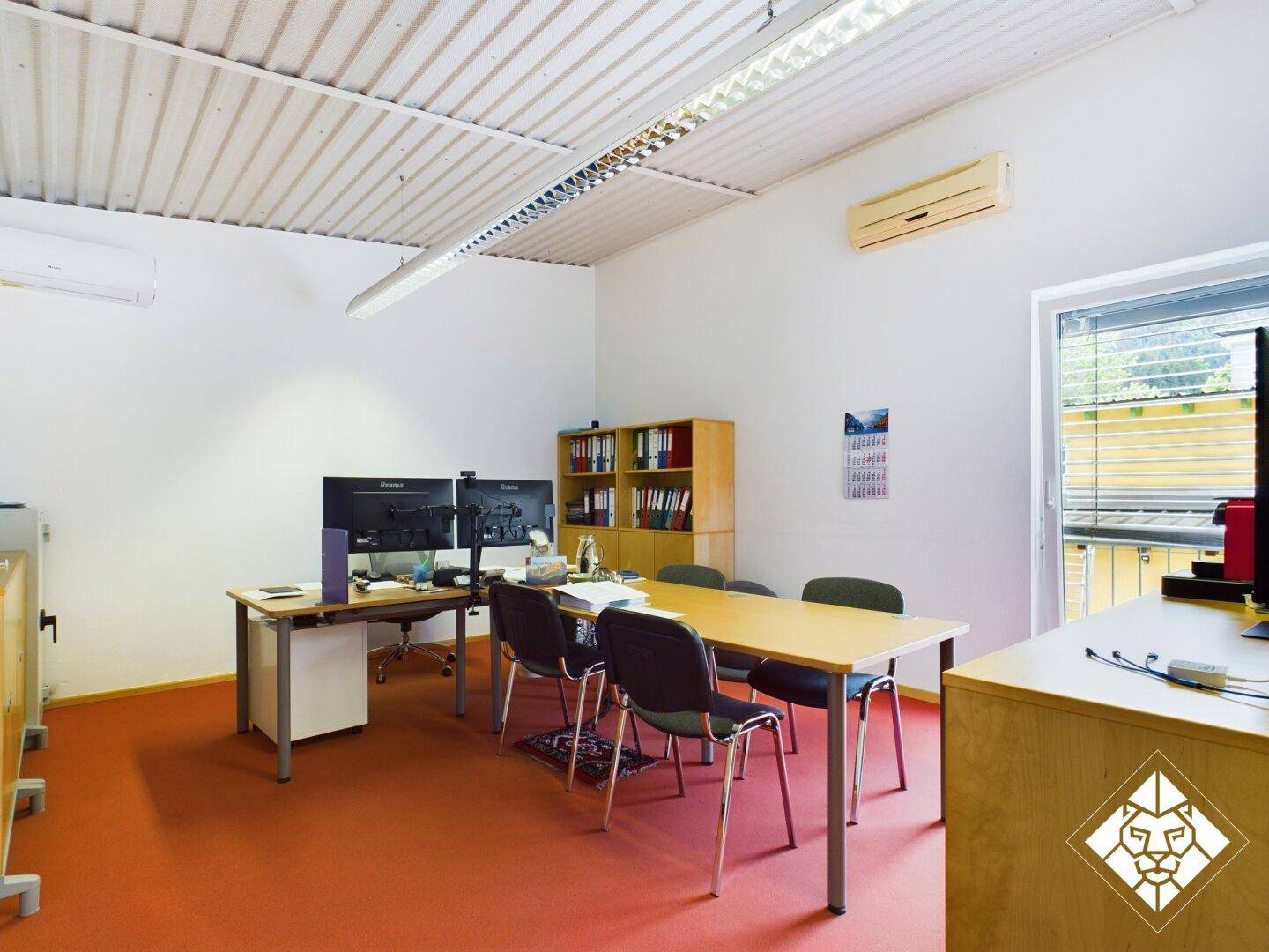 Helles Büro mit rotem Teppichboden, zwei Schreibtischen, Stühlen und einer Klimaanlage an der Wand.