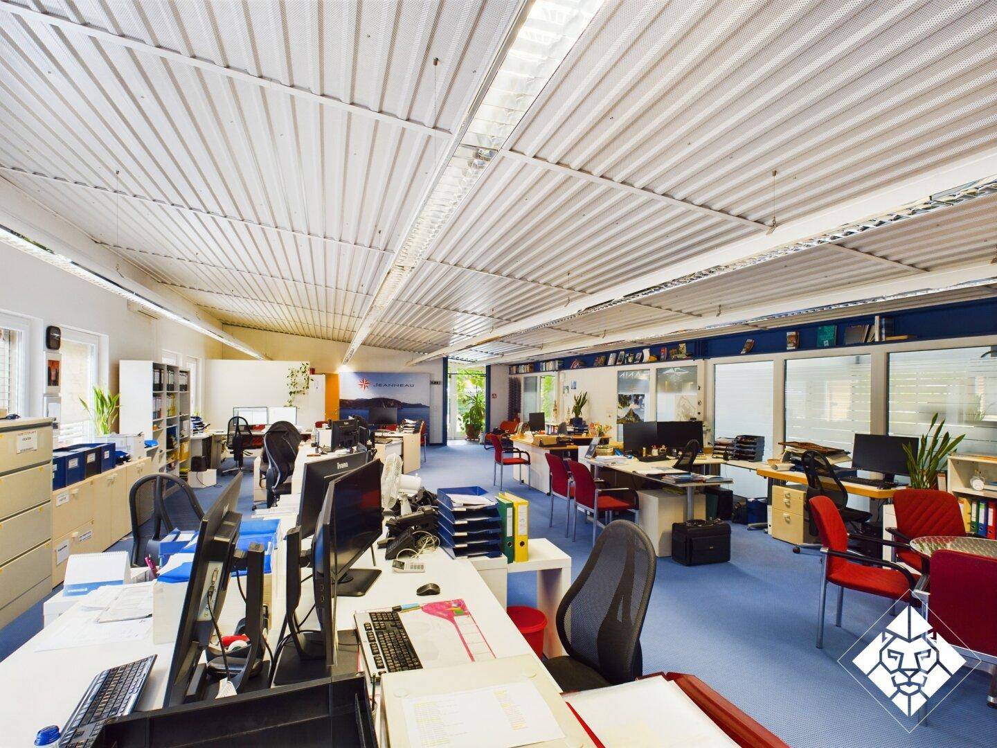Weitläufiges Büro mit vielen Schreibtischen, Computern und blauen Teppichböden unter einer Wellblechdecke.