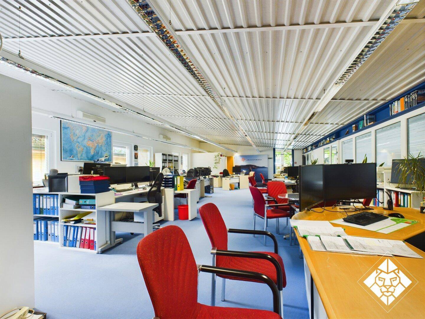 Großes Großraumbüro mit zahlreichen Arbeitsplätzen, blauen Teppichböden und abgehängter Decke.