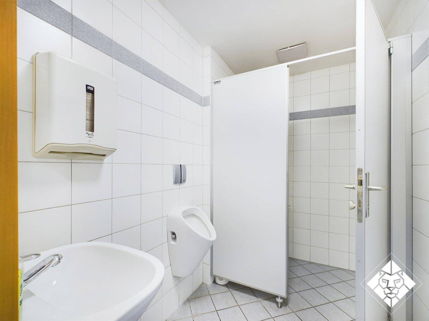 Helle, geflieste Herrentoilette mit Urinal, Waschbecken und Papierspender in einem Gewerbegebäude.