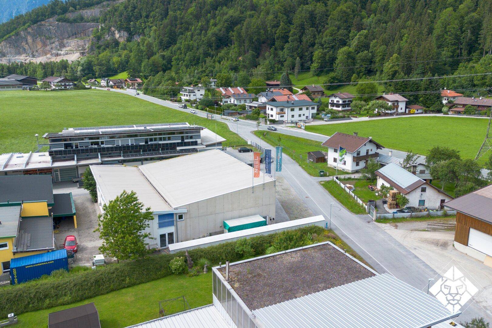 Luftaufnahme eines Gewerbegebäudes mit angrenzender Straße und Wohnhäusern vor einer Bergkulisse.