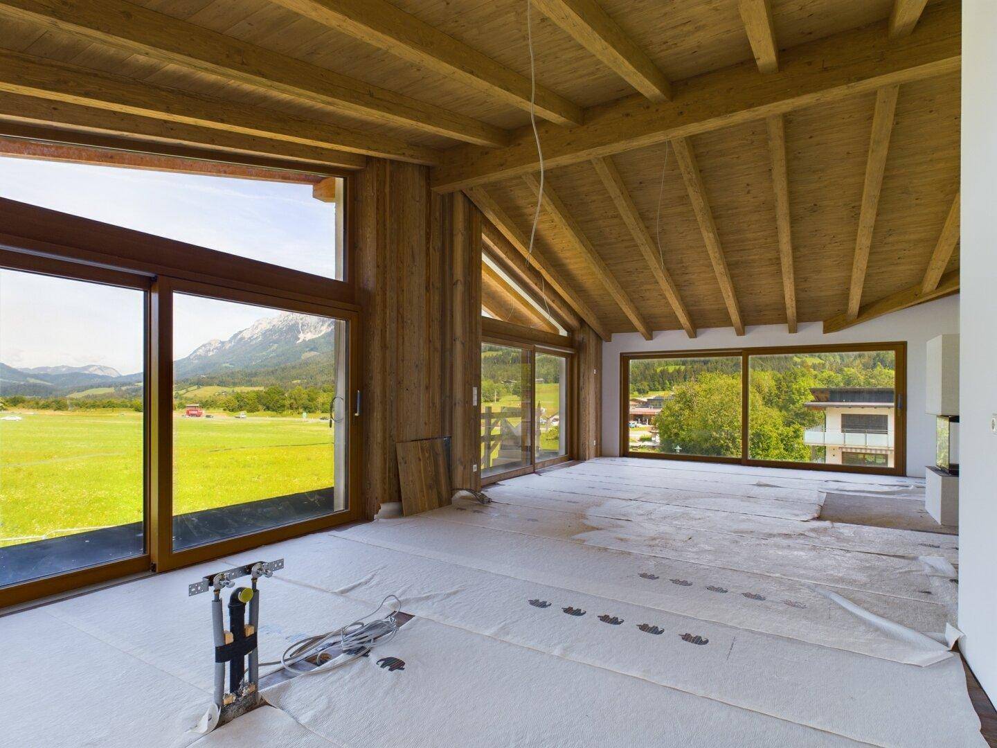 Geräumiges Wohnzimmer im Rohbau mit Holzbalkendecke, großen Fenstern und weitem Bergblick.