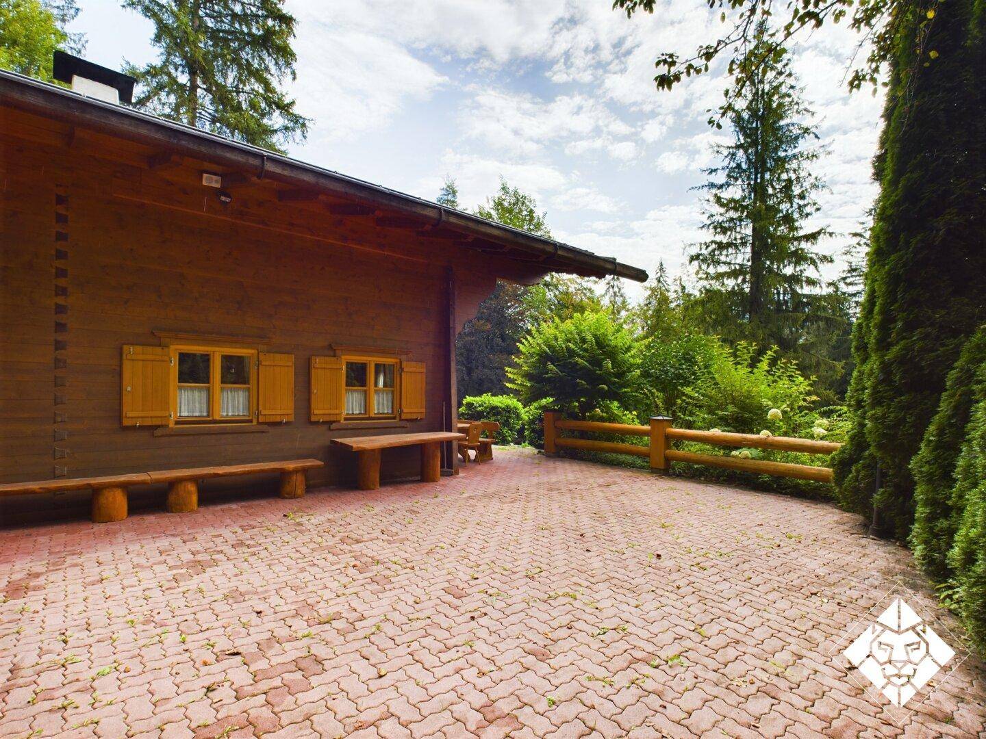 Große gepflasterte Fläche neben dem Holzhaus, umgeben von üppiger Vegetation und Bäumen.