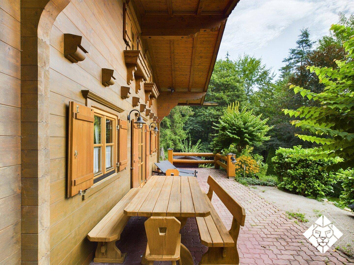 Rustikale Holzmöbel auf der gepflasterten Terrasse neben dem Holzhaus, umgeben von Natur.