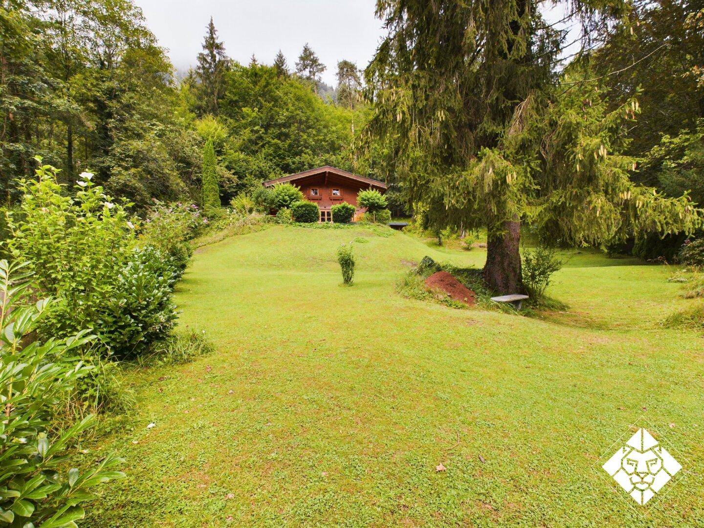 Charmantes Holzhaus auf einem Hügel, umgeben von einem weitläufigen grünen Garten und hohen Bäumen.