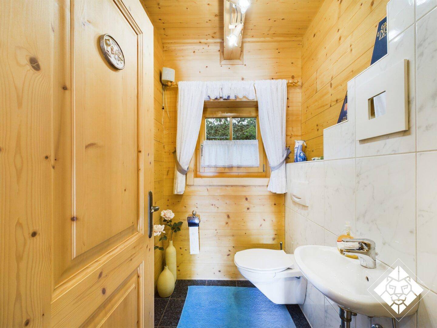 Kleines WC mit Holzvertäfelung, Fenster und weiß gefliestem Bereich um Toilette und Waschbecken.