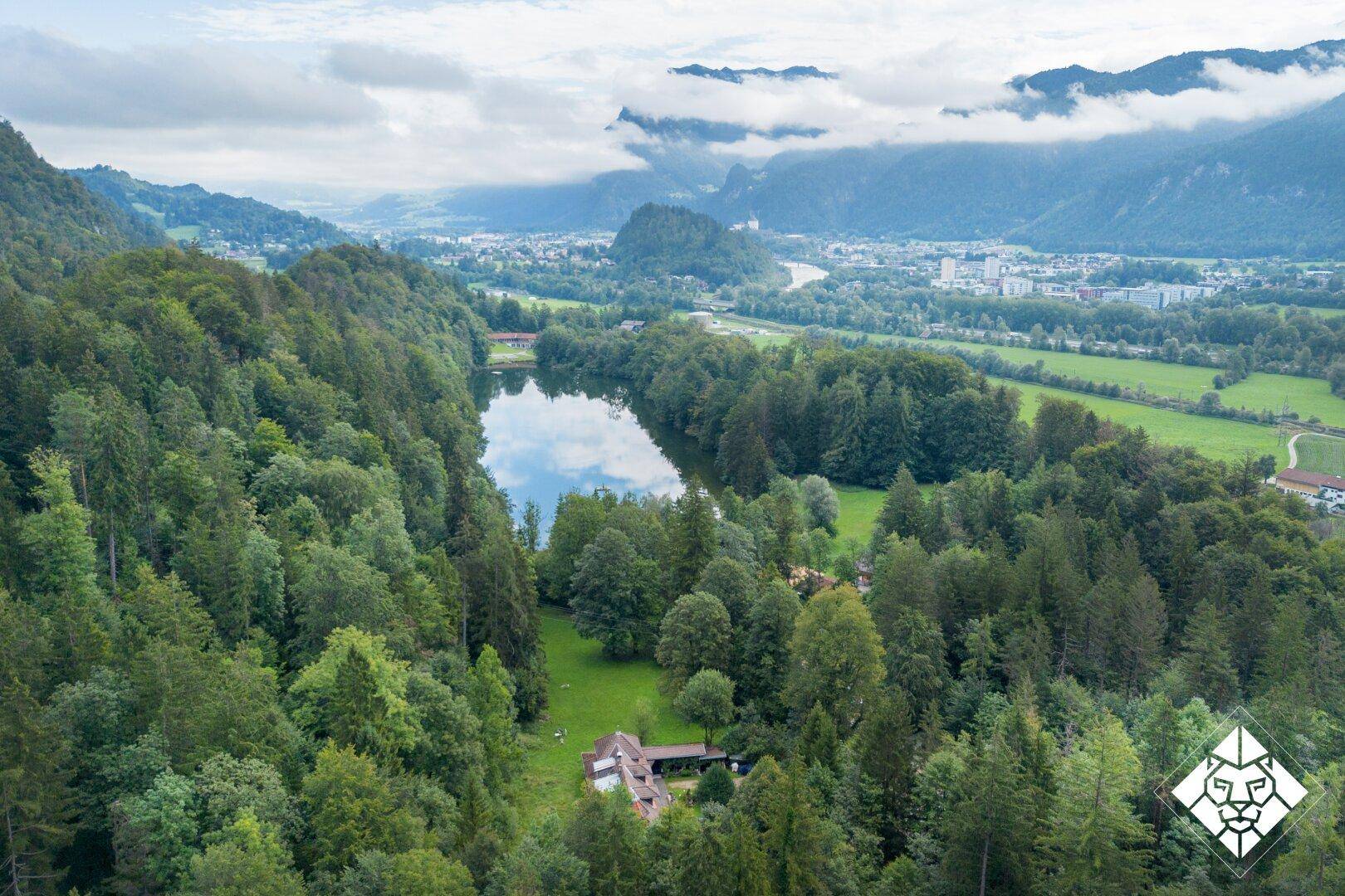 Panoramablick aus der Vogelperspektive auf ein Haus am See, umgeben von Wäldern und Bergen.