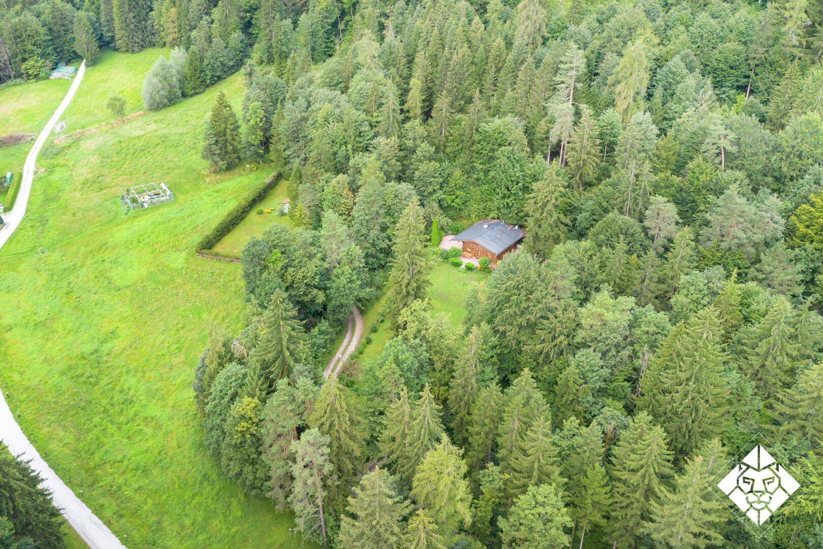 Weitläufige Luftaufnahme eines Holzhauses inmitten eines dichten Waldes und grüner Wiesen.