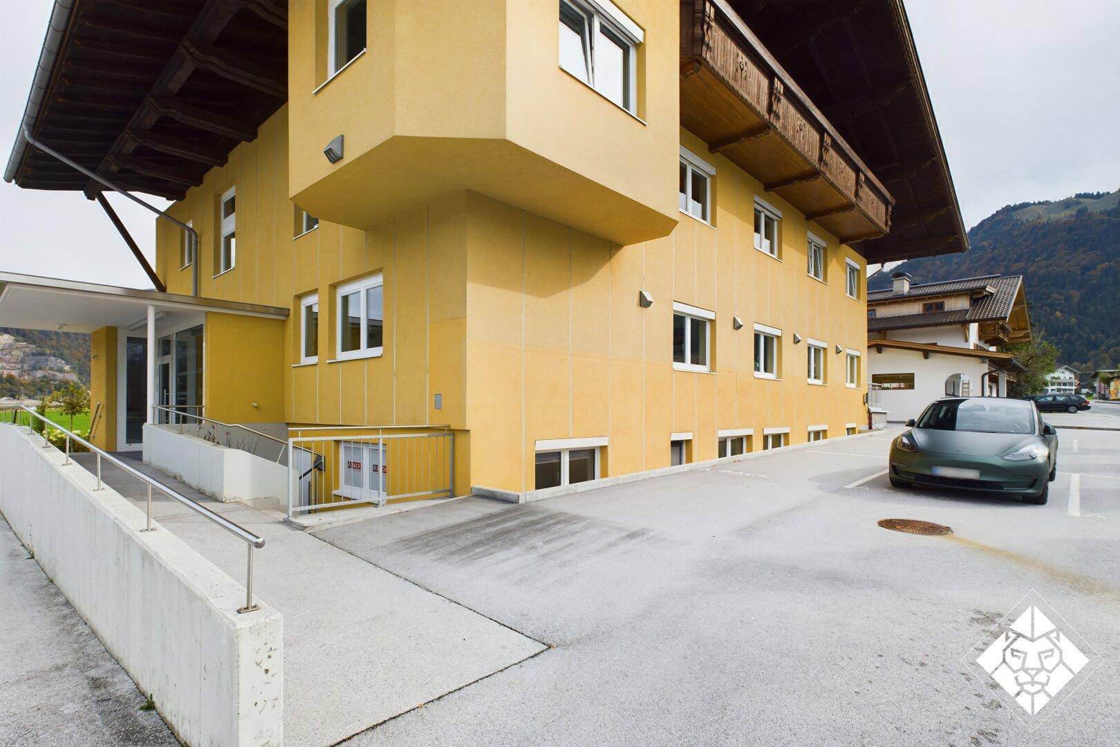 Gelbes Gebäude mit Parkplätzen und einer Rampe, umgeben von einer bergigen Landschaft.