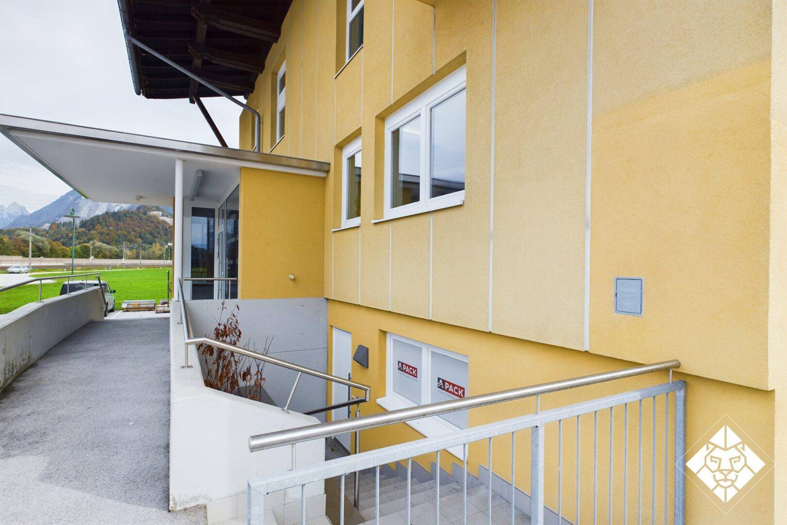 Gelbe Gebäudefassade mit Rampe und Treppe zum Eingangsbereich, umgeben von Bergen.