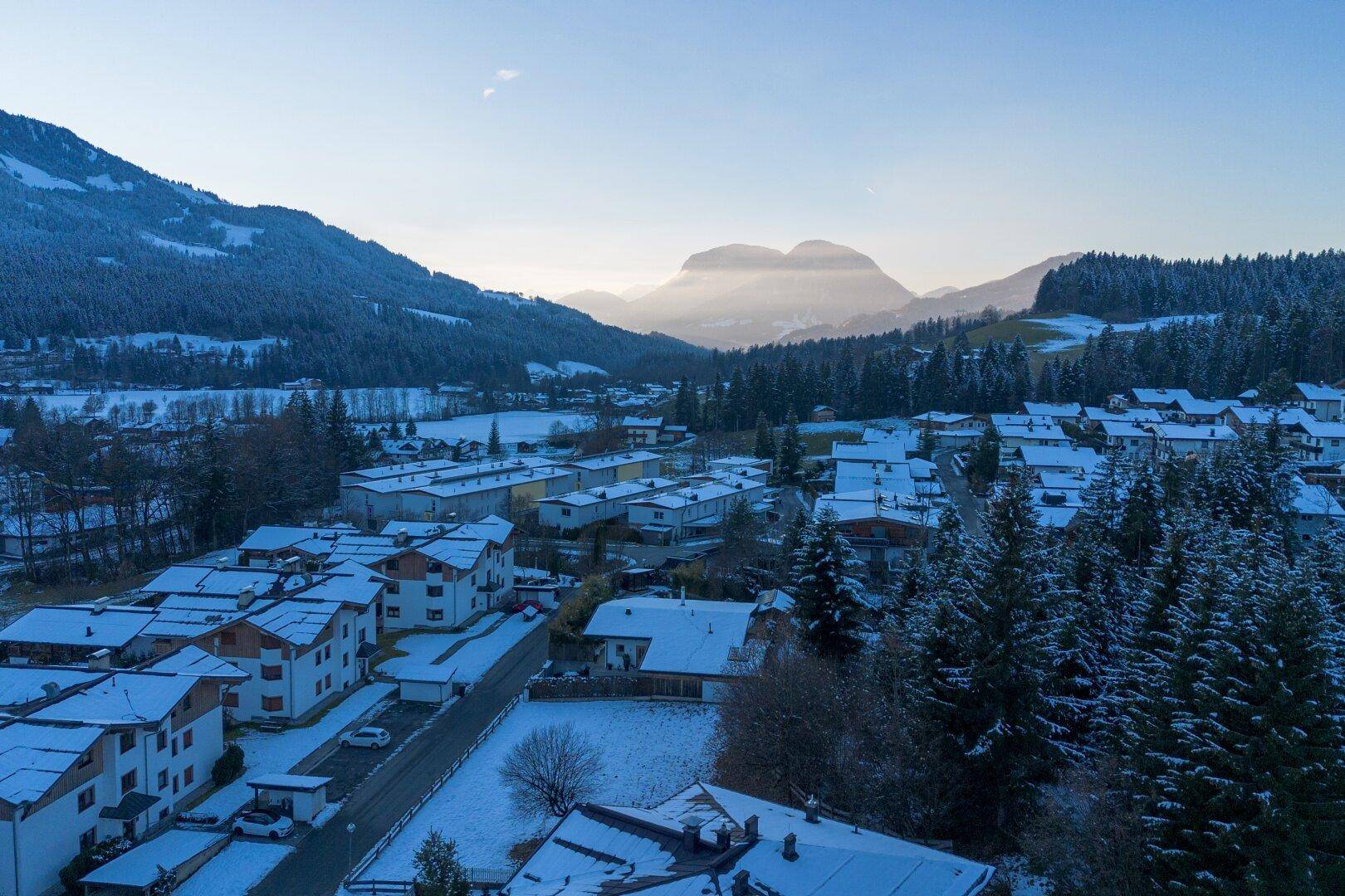 Panoramablick auf ein verschneites Dorf in einem Tal, umgeben von bewaldeten Bergen.