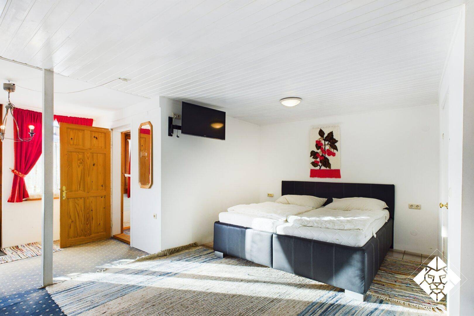 Helles Schlafzimmer mit Doppelbett, Teppichboden und Wandfernseher.
