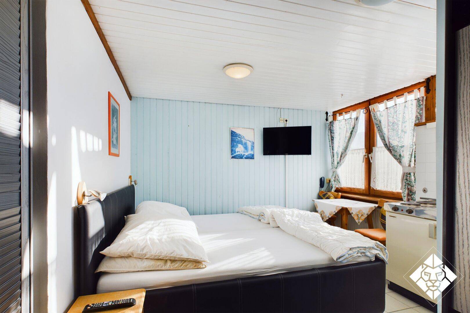 Schlafzimmer mit Doppelbett, kleiner Küchenzeile und Fenster mit Vorhängen.